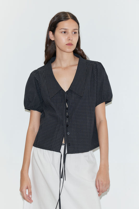 A woman with long brown hair wears the Fine Collar Top - Mono Grid by Deiji Studios, featuring a collared v-neck and short sleeves, paired with a white skirt against a plain white background.