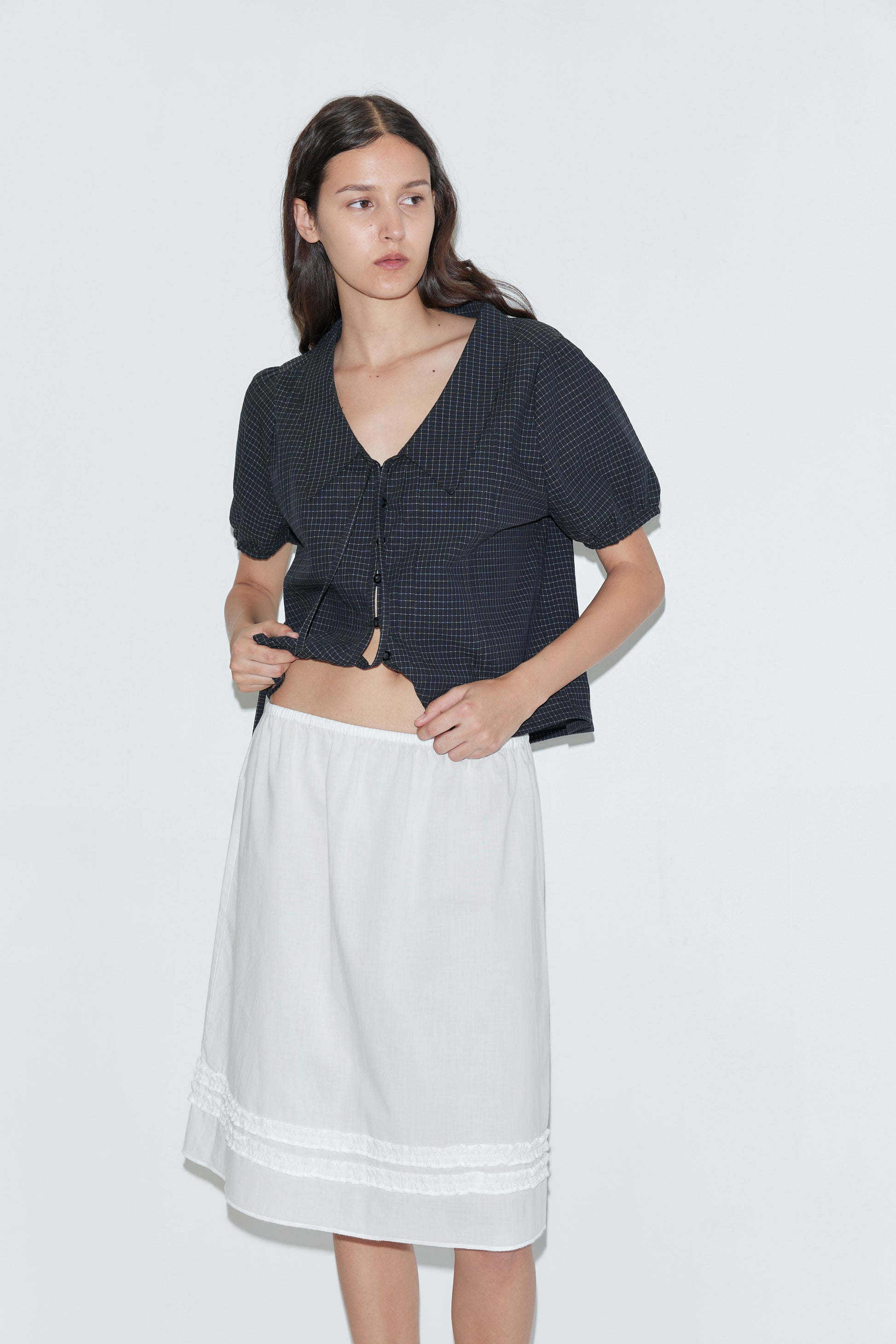 A woman with long dark hair wears a black short-sleeve blouse and the Daytime Skirt - White, a loose white cotton voile midi skirt with lace trim, standing against a plain light background.