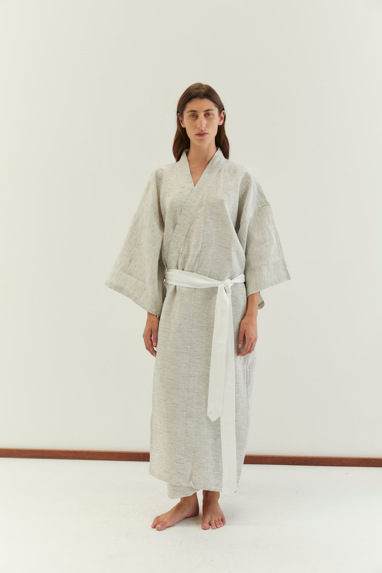 A person stands barefoot against a plain white background, wearing the 02 robe - pinstripe with wide sleeves, a white linen waist tie, and styled in a kimono fashion.
