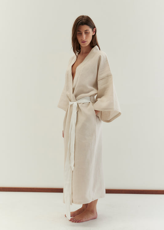 A woman with long brown hair and a calm expression stands barefoot against a white background, wearing the 02 robe - oatmeal. The full-length robe features wide sleeves and a linen waist tie.