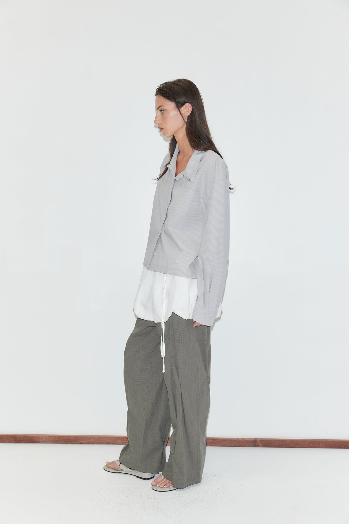 A woman with long dark hair stands in profile against a plain white background wearing the Boxy Shirt - Cloud Stripe by Deiji Studios, a pale grey-blue fine-striped boxy shirt, over a white top with olive wide-leg trousers and grey flip-flops.