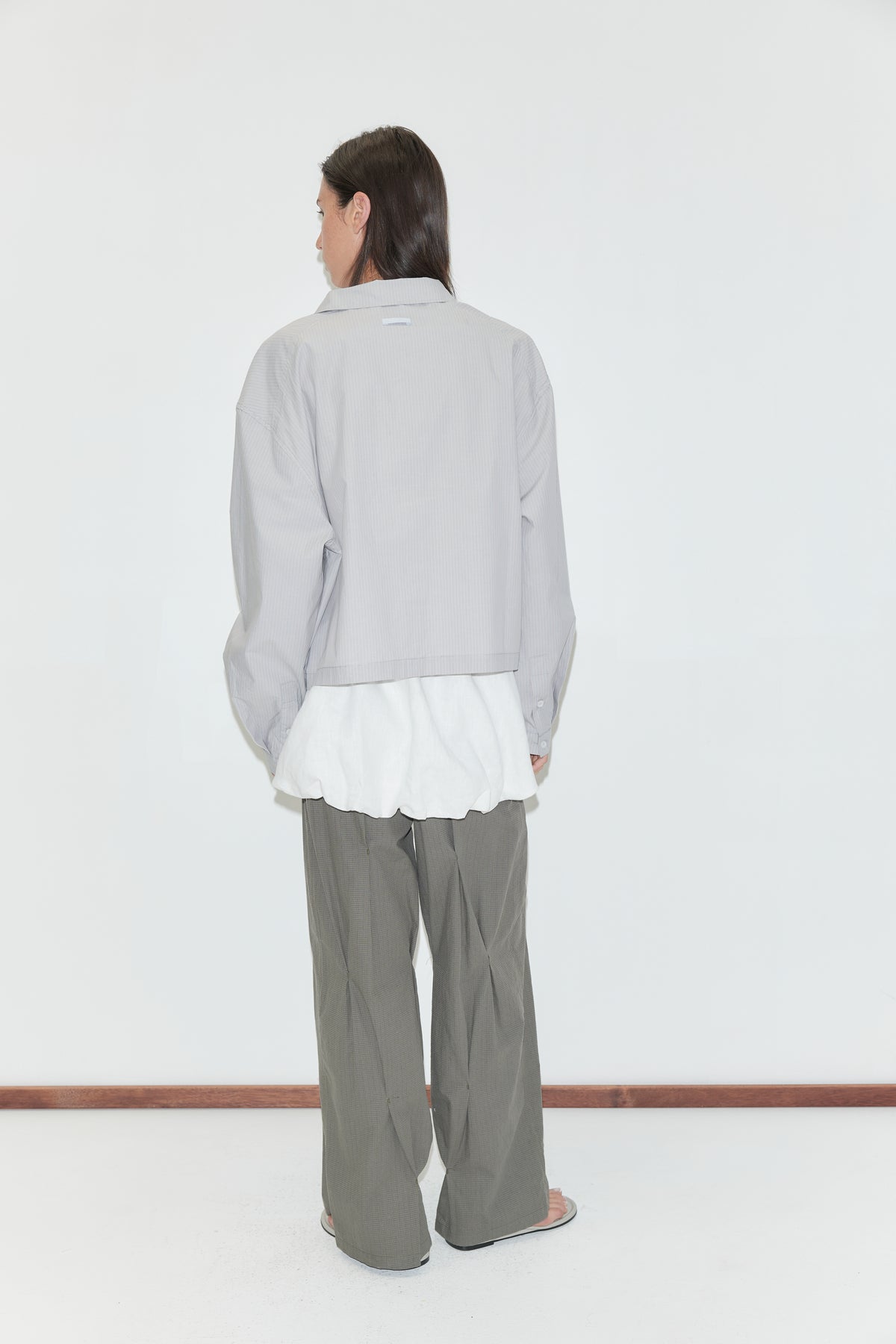 A woman with long dark hair stands with her back to the camera against a plain white background wearing the Boxy Shirt - Cloud Stripe by Deiji Studios, a pale grey-blue fine-striped boxy shirt, over a white top with olive wide-leg trousers.