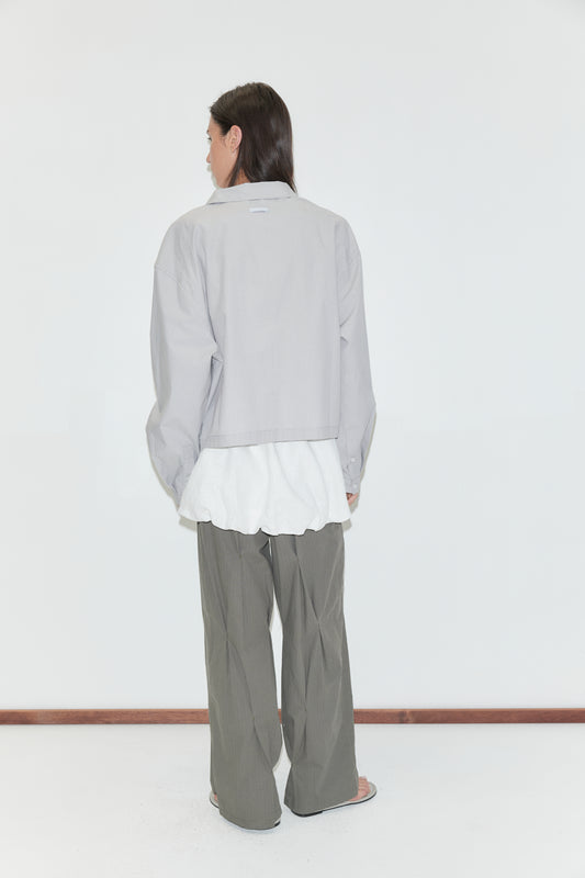 A woman with long dark hair stands with her back to the camera against a plain white background wearing the Boxy Shirt - Cloud Stripe by Deiji Studios, a pale grey-blue fine-striped boxy shirt, over a white top with olive wide-leg trousers.
