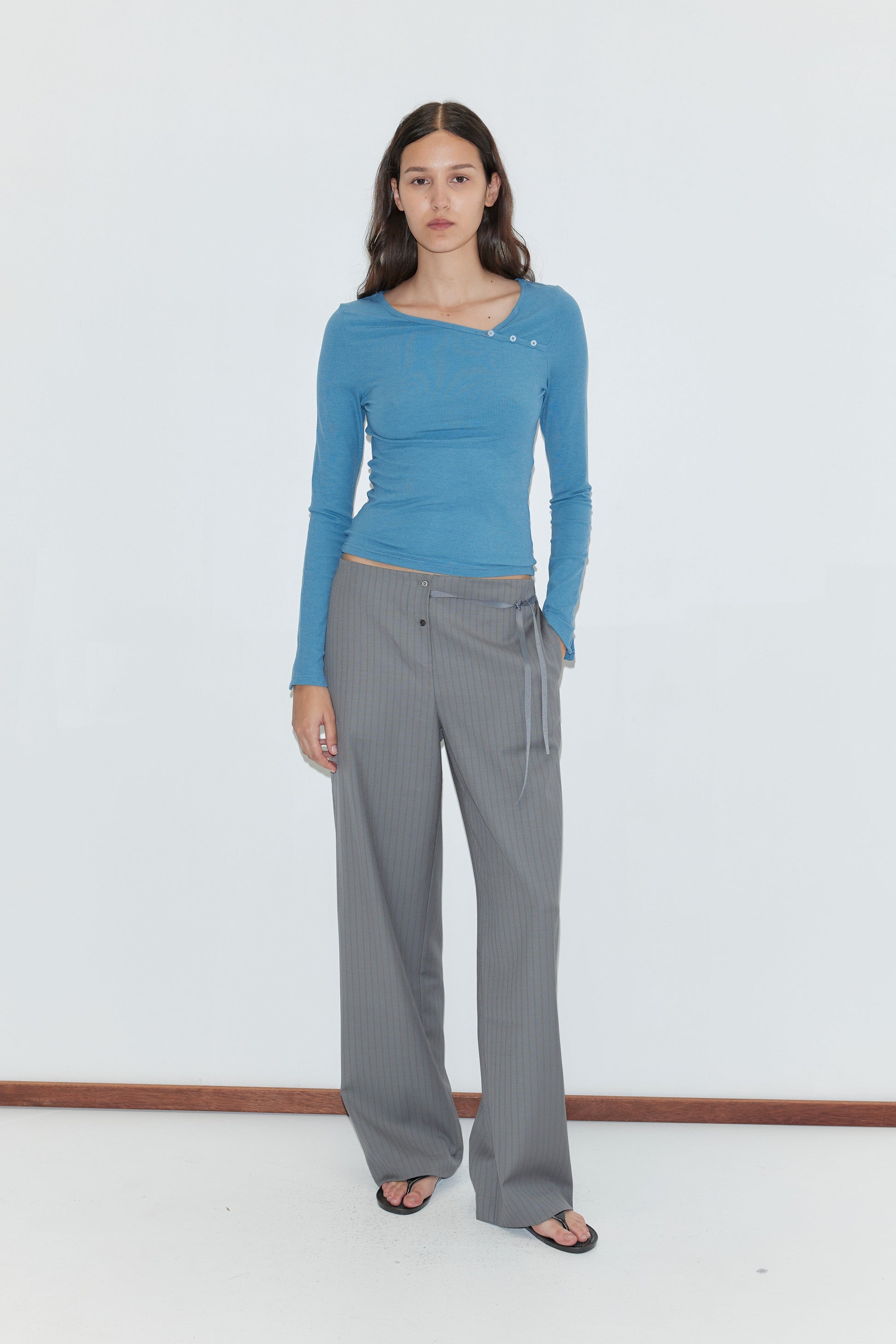 A person with long brown hair wearing a fitted blue asymmetric long-sleeve top and the Boxer Pant - Grey Stripe, featuring a relaxed elasticated waistband, stands indoors against a plain white wall, paired with black sandals.