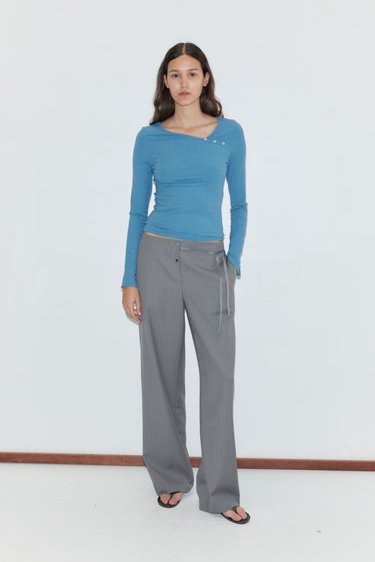 A person with long brown hair wearing a fitted blue asymmetric long-sleeve top and the Boxer Pant - Grey Stripe, featuring a relaxed elasticated waistband, stands indoors against a plain white wall, paired with black sandals.
