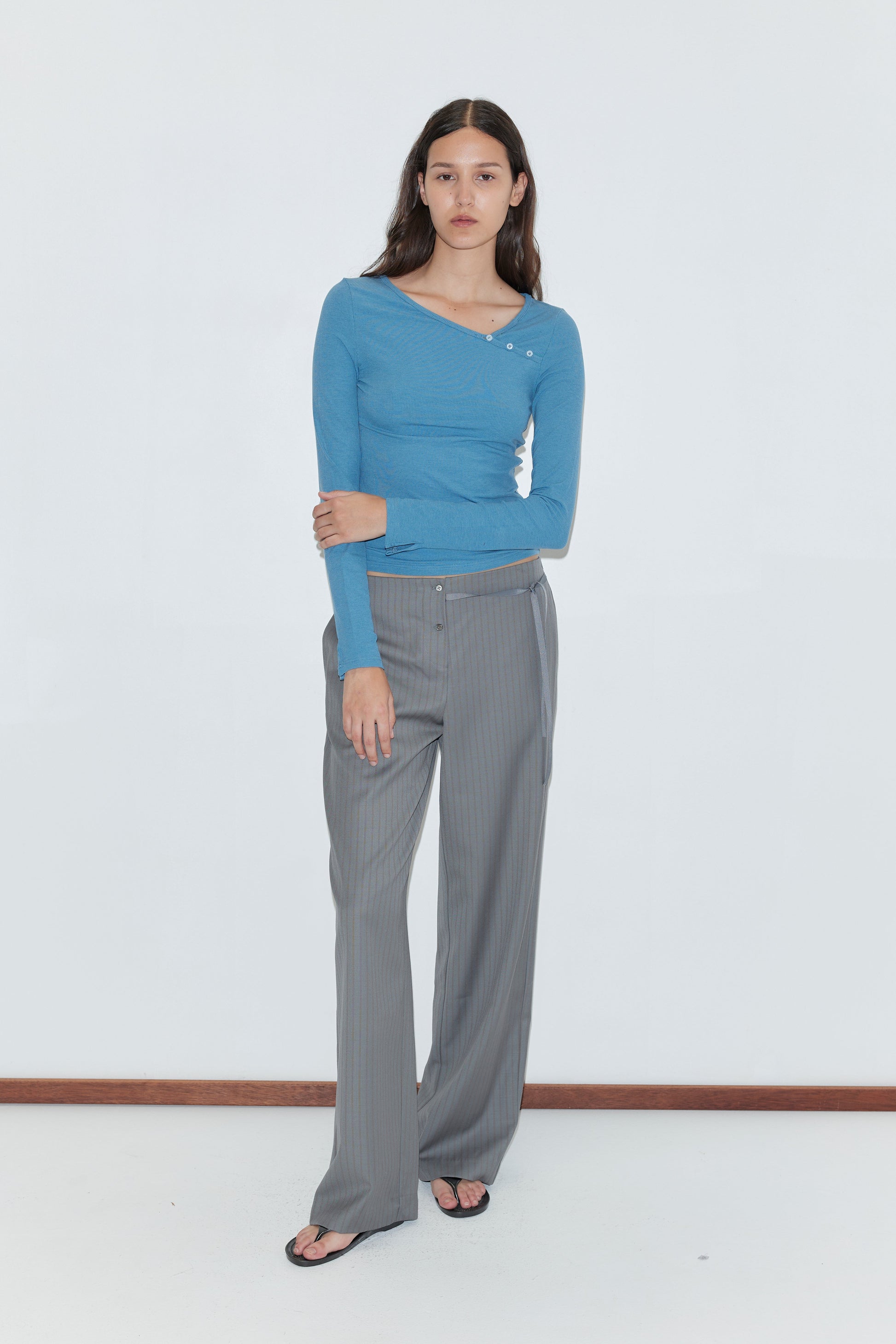A woman with long dark hair stands against a white background, wearing the Deiji Studios Multi Button Top in Aqua with gray wide-leg trousers and black flip-flops. The top features long sleeves and an asymmetric neckline.