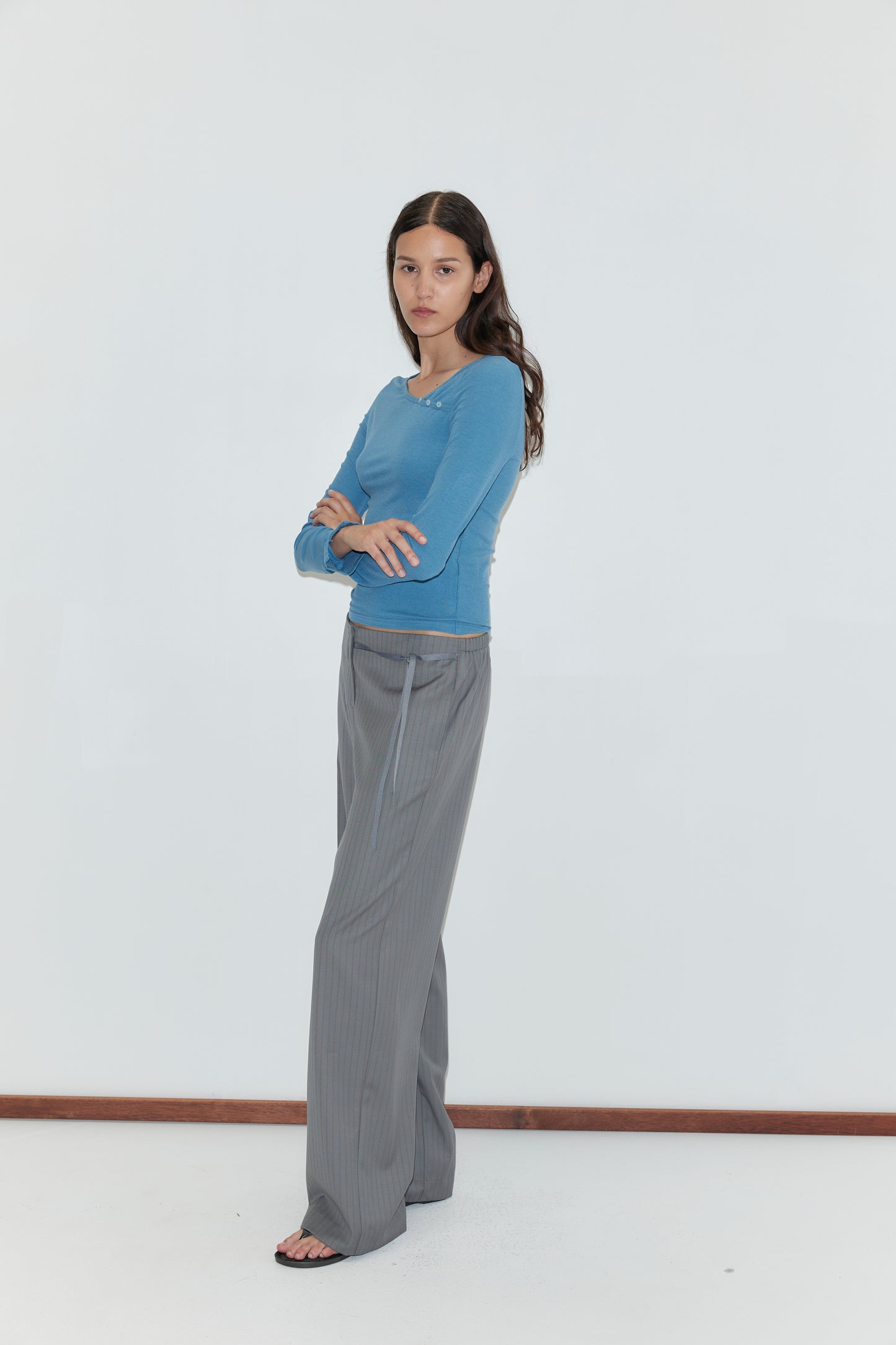 A woman with long brown hair stands against a plain white background, wearing the Multi Button Top in Aqua, paired with gray wide-leg pants and black sandals. She has her arms crossed and looks directly at the camera.