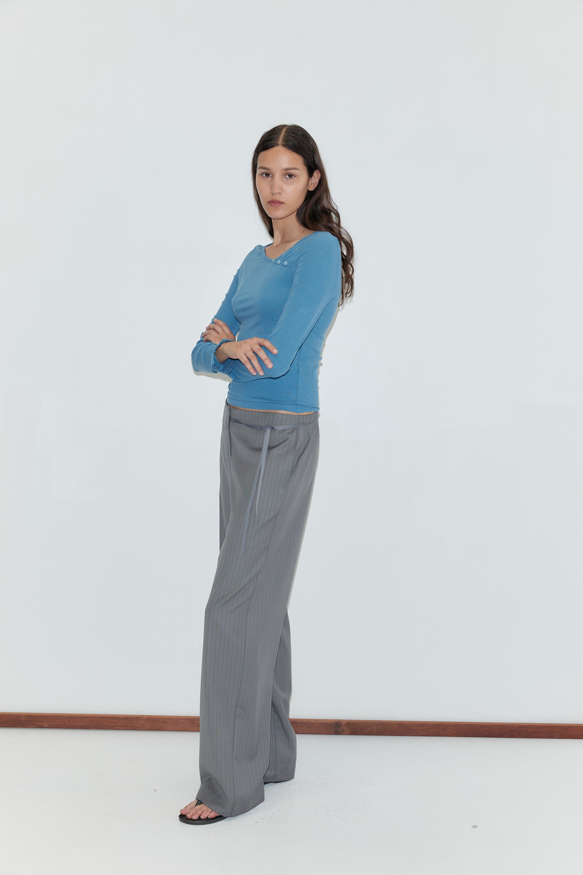 A woman with long brown hair stands against a plain white background, wearing the Multi Button Top in Aqua, paired with gray wide-leg pants and black sandals. She has her arms crossed and looks directly at the camera.