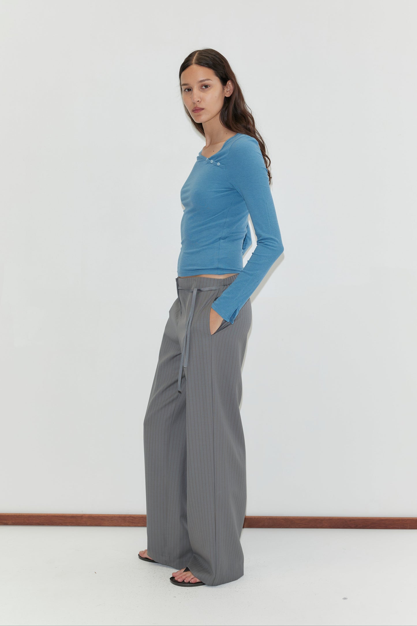 A woman with long brown hair stands against a white background, wearing the Deiji Studios Multi Button Top in Aqua, gray wide-leg drawstring trousers, and black sandals. She has her hands in her pockets and faces slightly sideways.