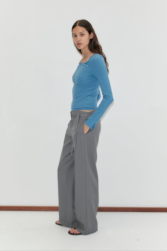 A woman with long brown hair stands against a white background, wearing the Deiji Studios Multi Button Top in Aqua, gray wide-leg drawstring trousers, and black sandals. She has her hands in her pockets and faces slightly sideways.