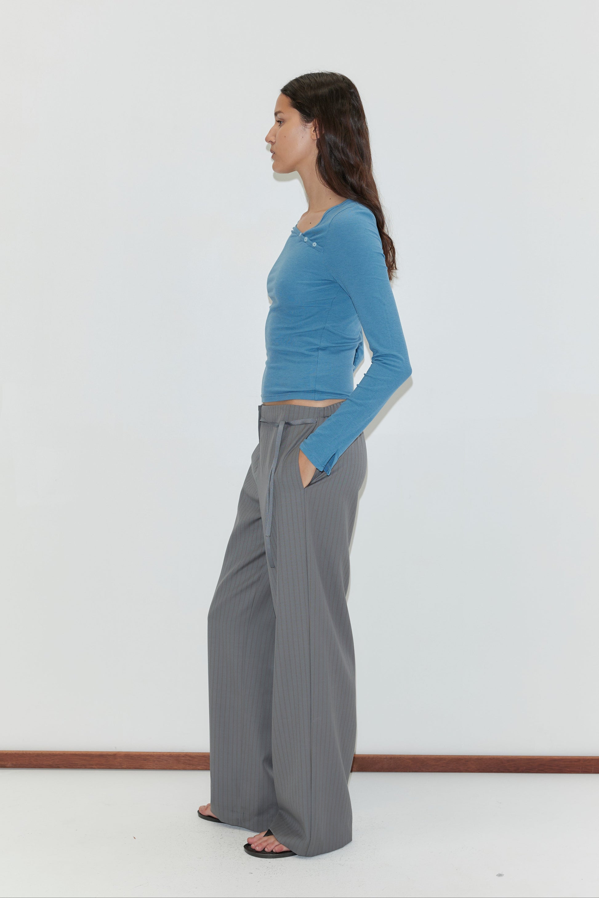 A woman stands in profile against a white background, wearing the Boxer Pant - Grey Stripe with a relaxed elasticated waistband, blue long-sleeve top, and black sandals. Her hands are in her pockets and she has long dark hair.