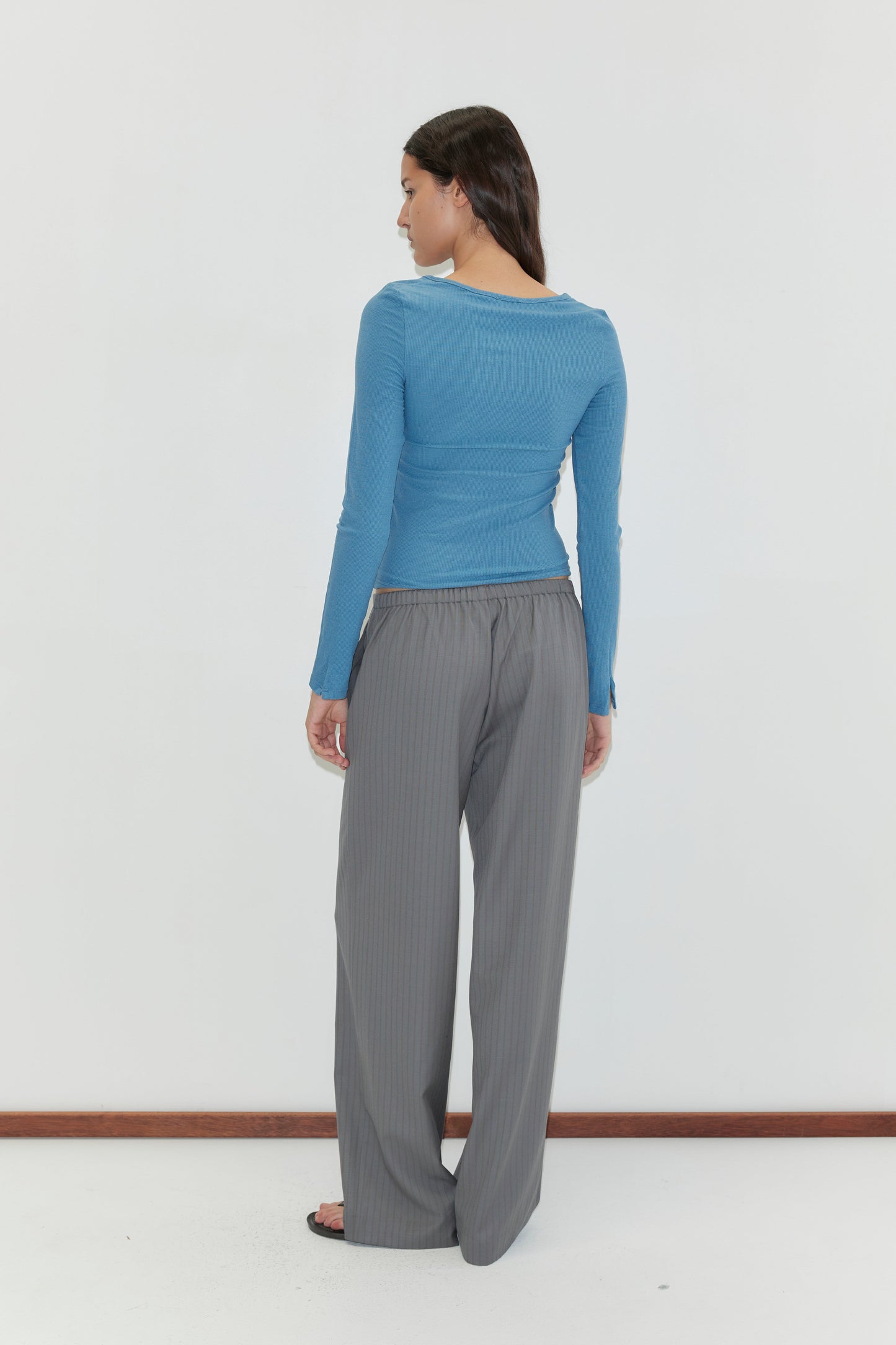A person with long dark hair, wearing a blue long-sleeve top and the Boxer Pant - Grey Stripe, stands barefoot facing away in front of a plain white wall.