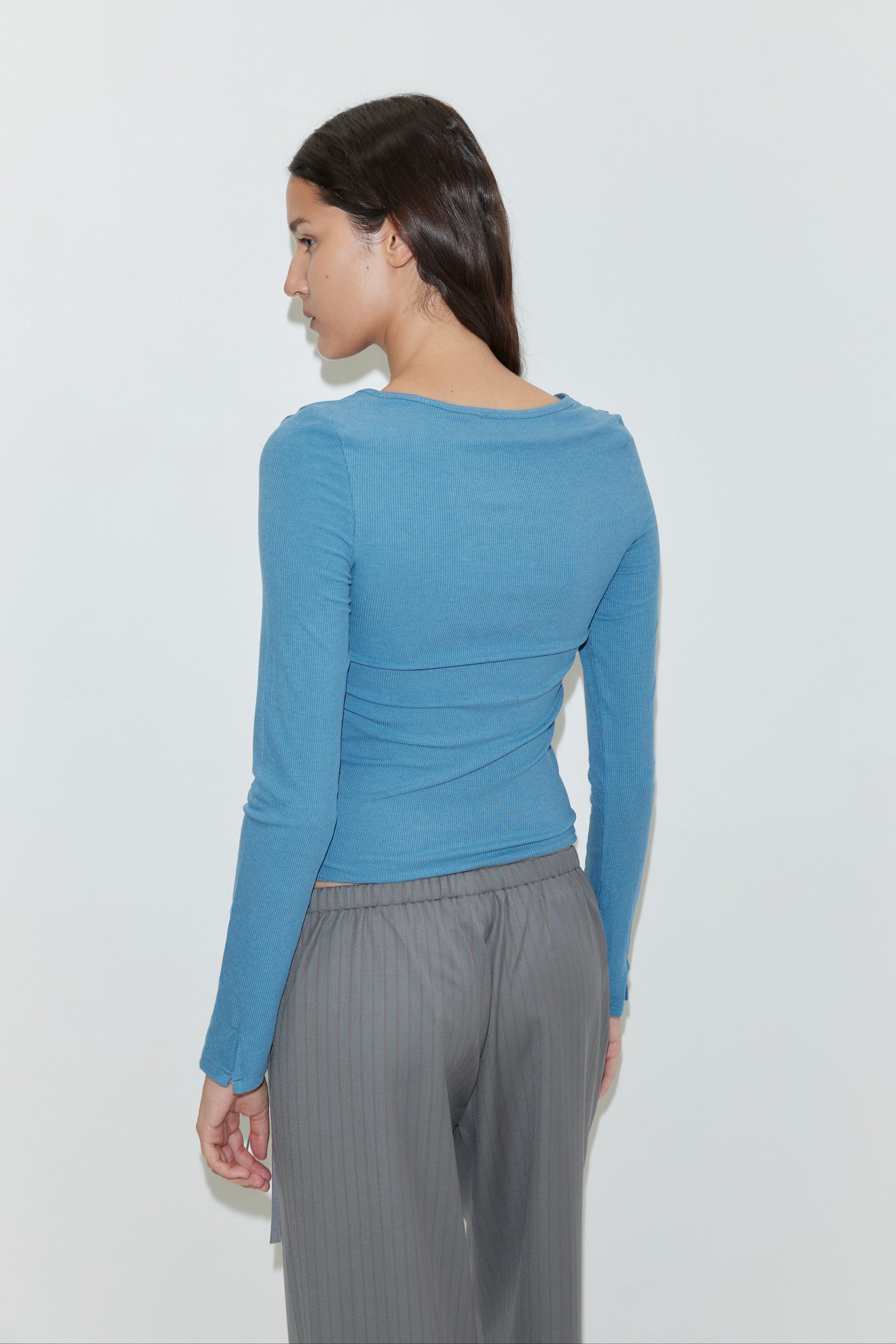 A woman with long brown hair, seen from behind, wears the Deiji Studios Multi Button Top in Aqua and loose gray pants, set against a plain light background.