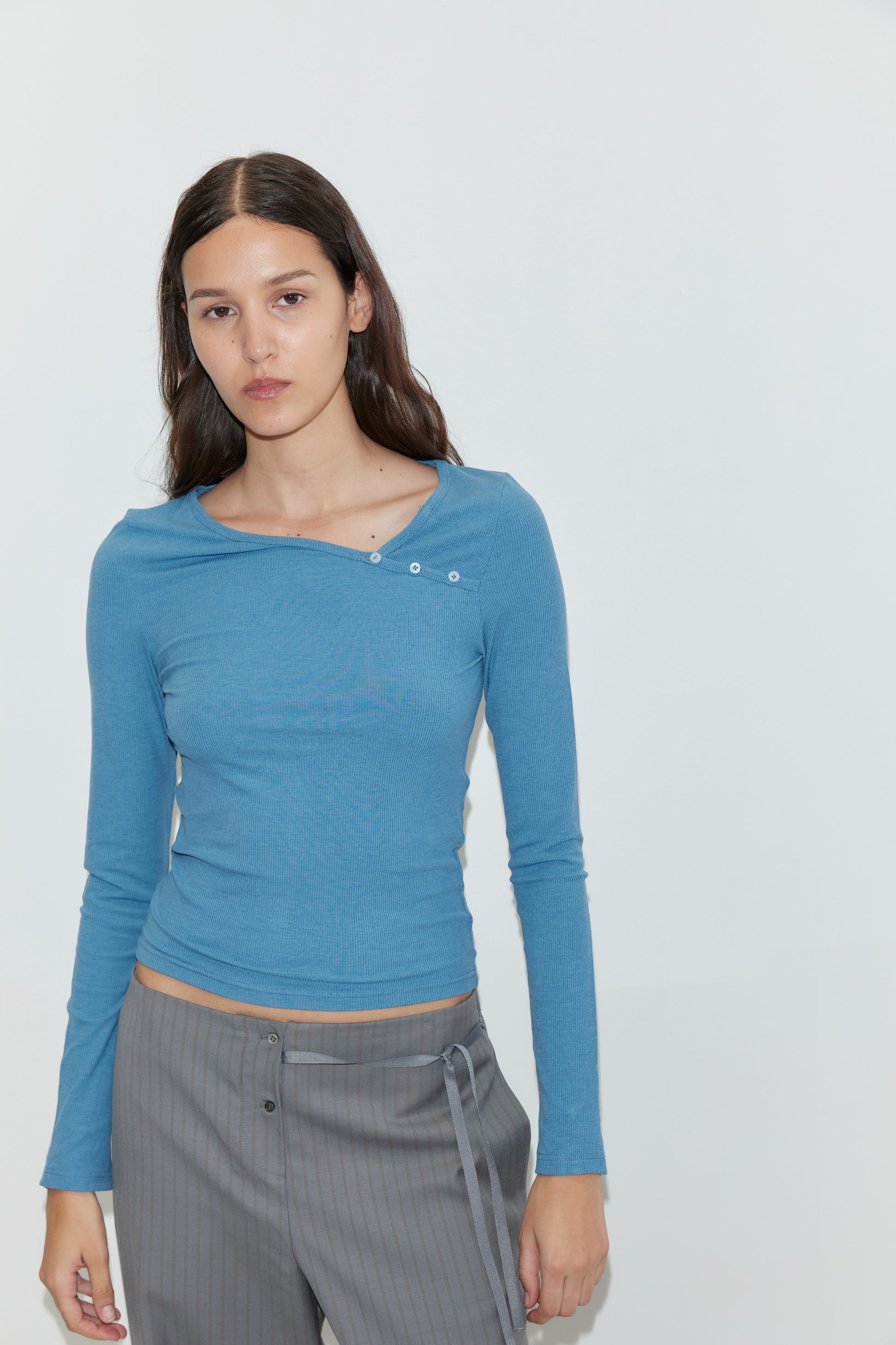 A woman with long brown hair wears the Multi Button Top in Aqua, a fitted long sleeve jersey featuring an asymmetric scoop neckline and decorative buttons, paired with gray striped pants as she stands against a plain white background.