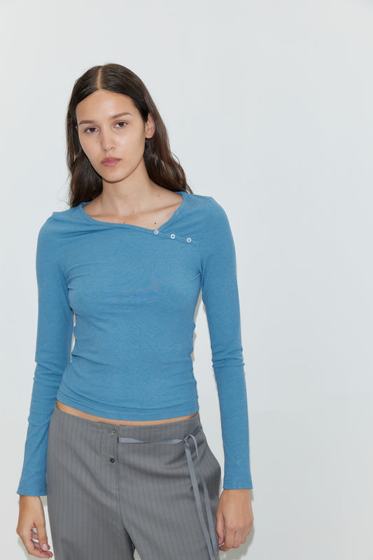 A woman with long brown hair wears the Multi Button Top in Aqua, a fitted long sleeve jersey featuring an asymmetric scoop neckline and decorative buttons, paired with gray striped pants as she stands against a plain white background.