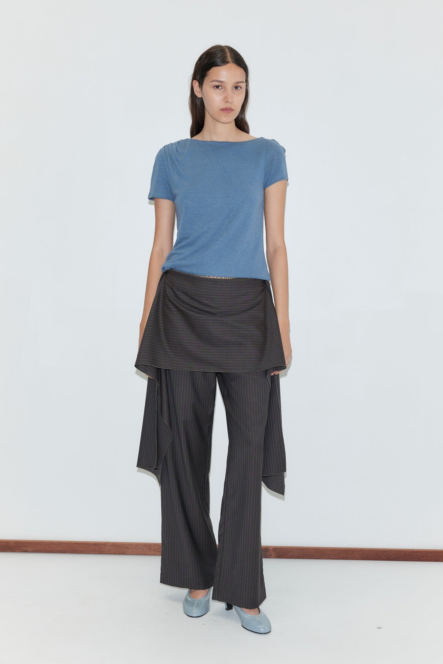 A woman with long dark hair and a neutral expression stands against a white background, wearing the Drape Tee - Blue by Deiji Studios, gray and black pinstripe pants with hip draping, and light blue heels.