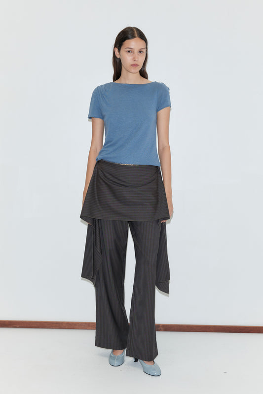 A woman with long dark hair and a neutral expression stands against a white background, wearing the Drape Tee - Blue by Deiji Studios, gray and black pinstripe pants with hip draping, and light blue heels.