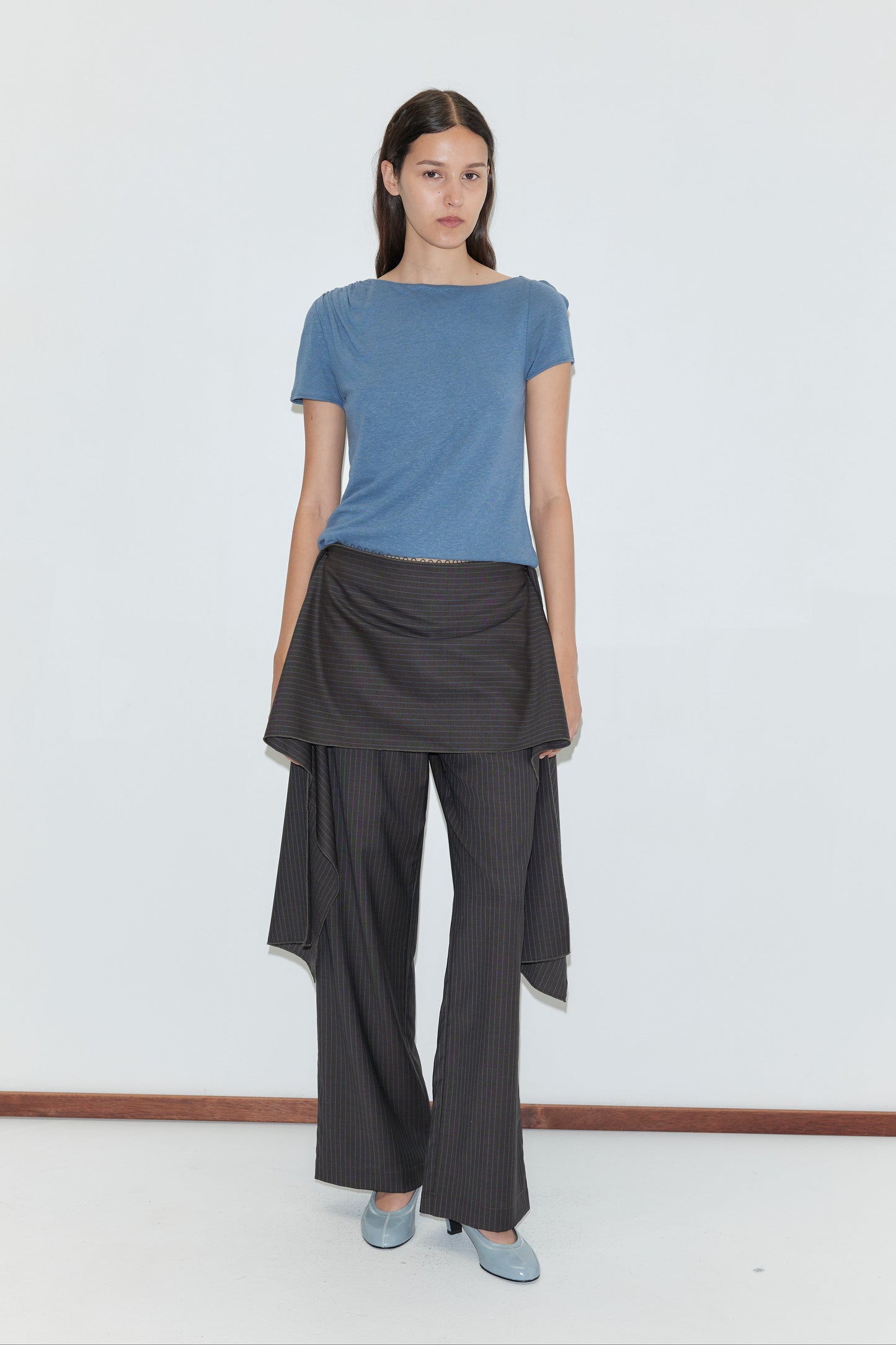 A woman with long brown hair wears a blue t-shirt, wide-legged pinstriped pants with an asymmetrical overlay, light blue shoes, and the Pure Wool Belt - Charcoal Stripe from Deiji Studios, standing against a plain white background.