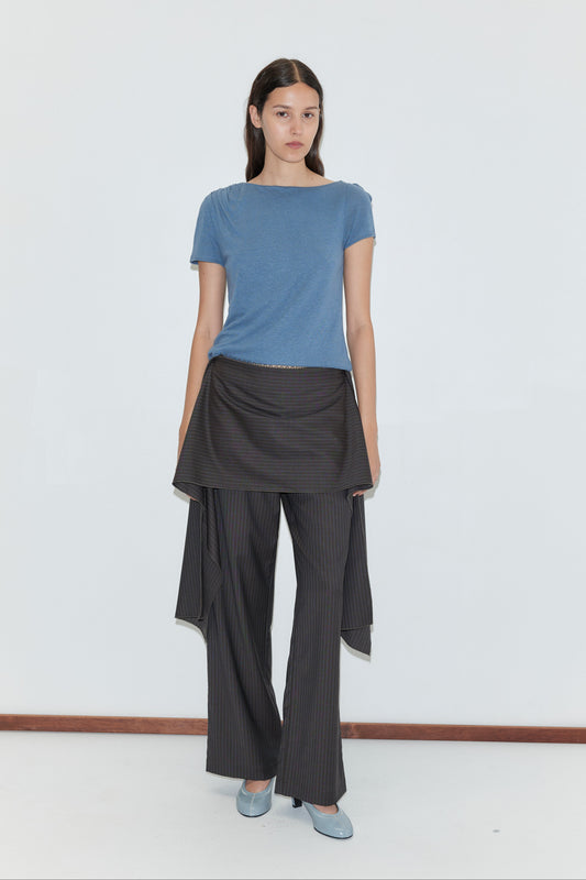 A woman with long brown hair wears a blue t-shirt, wide-legged pinstriped pants with an asymmetrical overlay, light blue shoes, and the Pure Wool Belt - Charcoal Stripe from Deiji Studios, standing against a plain white background.