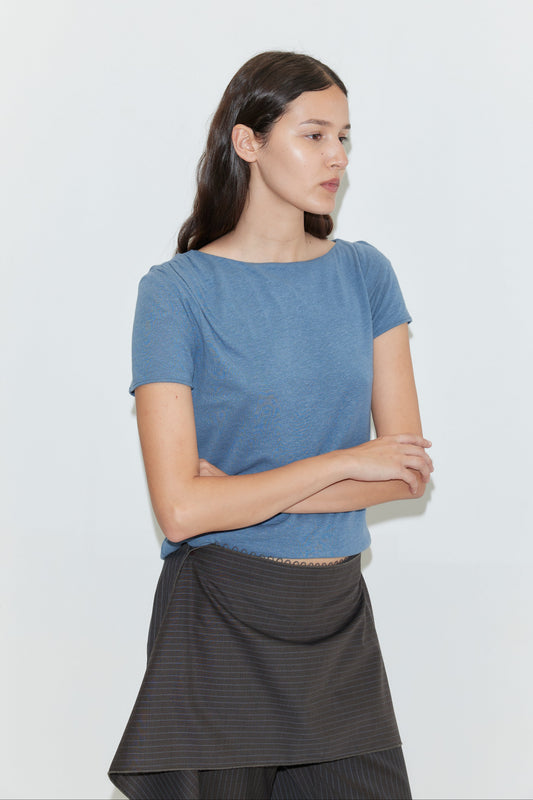 A woman with long dark hair stands against a plain white background, wearing the Drape Tee - Blue with a dark skirt. Her arms are gently crossed as she looks off to the side.