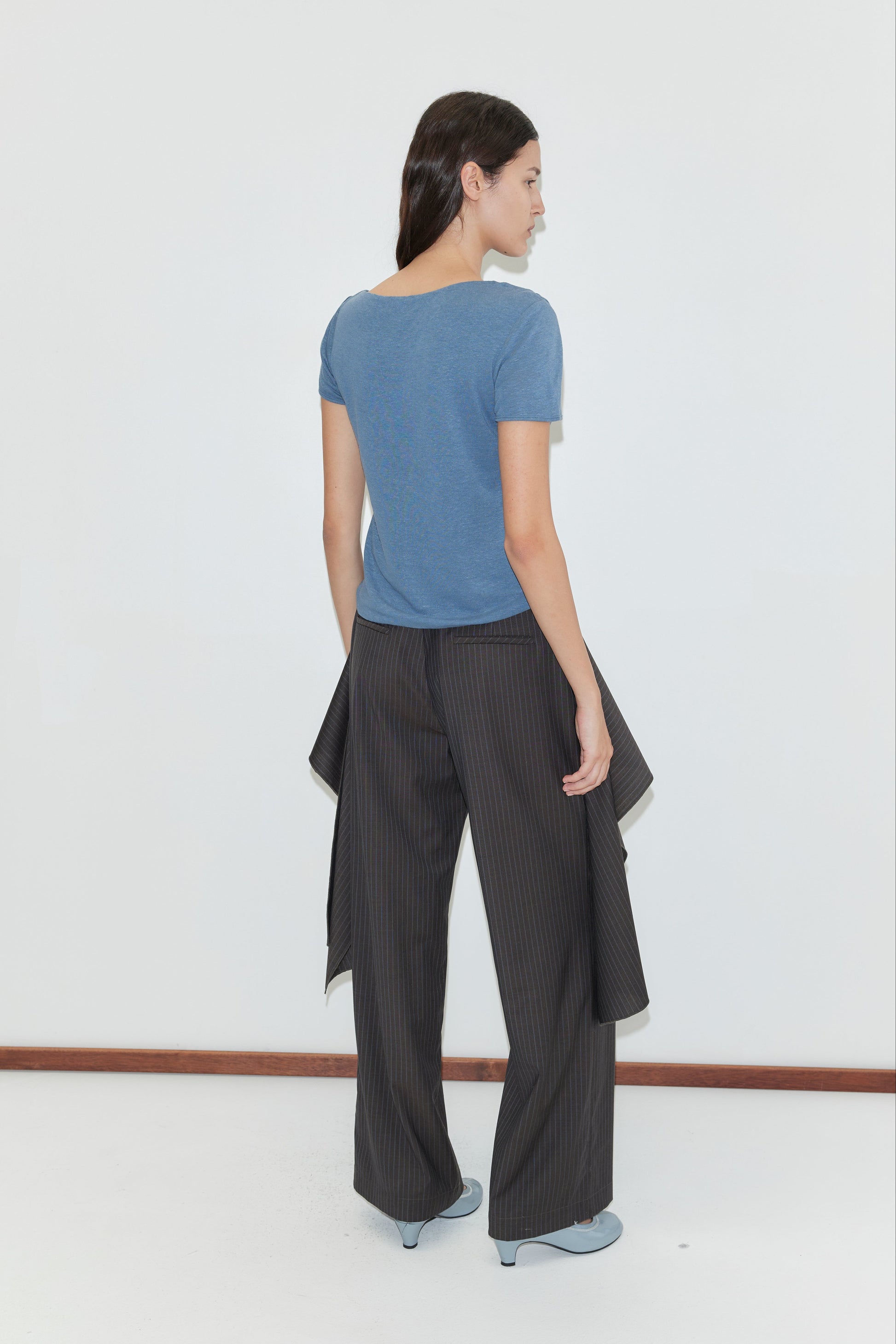A woman faces away from the camera, wearing the Drape Tee - Blue by deiji studios, wide-leg pinstriped pants with fabric panels, and light blue shoes, standing against a plain white wall.