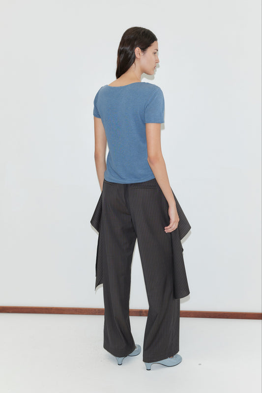 A woman faces away from the camera, wearing the Drape Tee - Blue by deiji studios, wide-leg pinstriped pants with fabric panels, and light blue shoes, standing against a plain white wall.