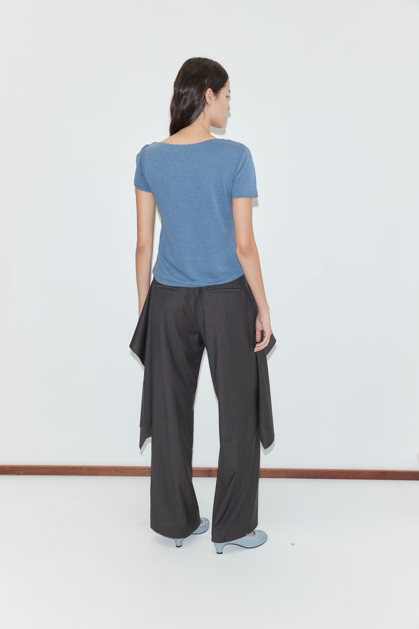 A woman stands facing away against a white background, wearing a blue short-sleeve top, blue heels, and the Pure Wool Zip Pant - Charcoal Stripe with relaxed legs and draped detail.
