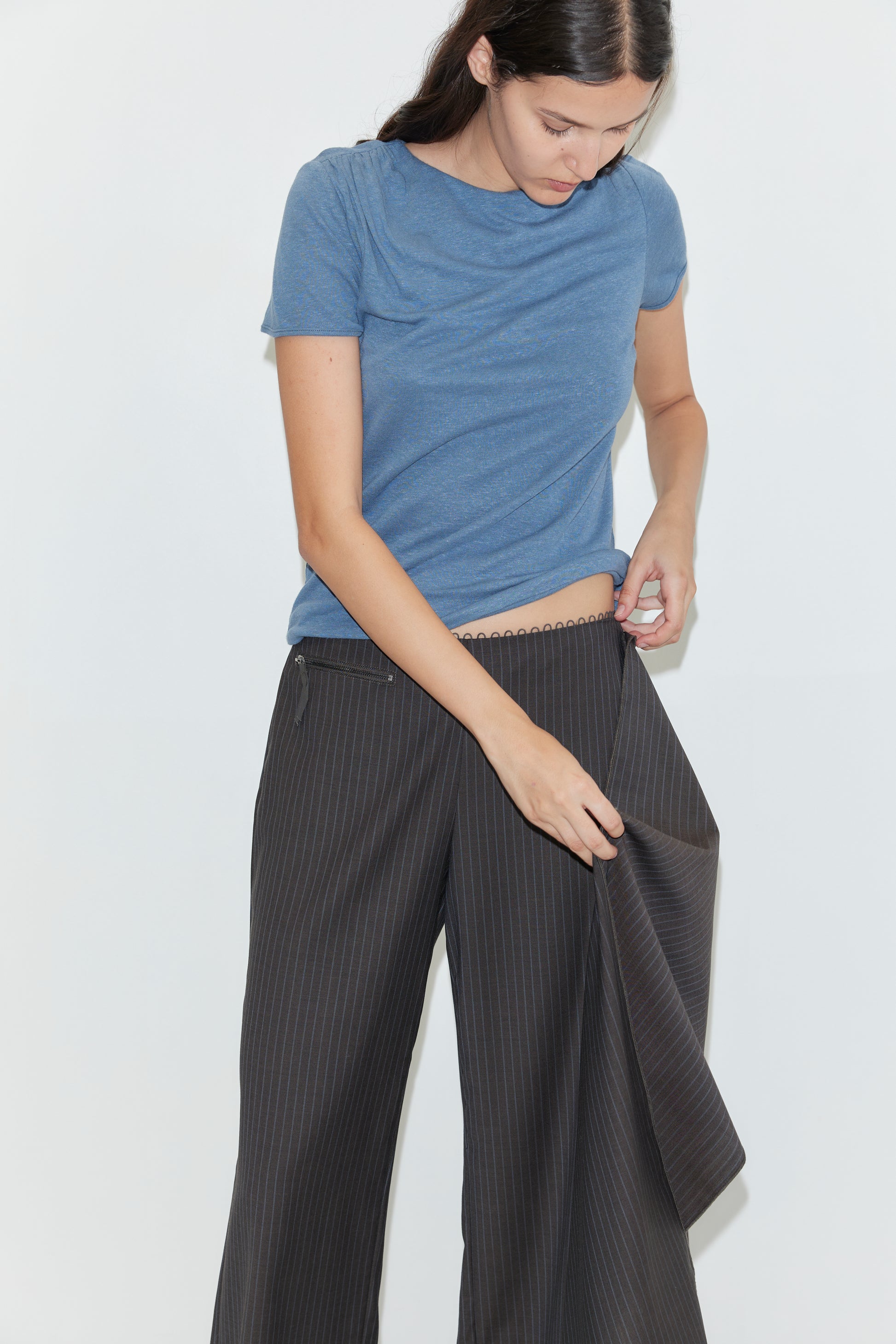 A woman with long dark hair, wearing a blue t-shirt and dark pinstriped wide-leg pants, adjusts the Pure Wool Belt - Charcoal Stripe at her waistband against a plain white background.