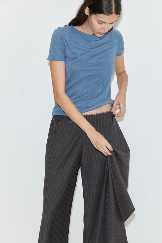 A woman with long dark hair, wearing a blue t-shirt and dark pinstriped wide-leg pants, adjusts the Pure Wool Belt - Charcoal Stripe at her waistband against a plain white background.