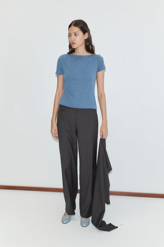 A woman stands against a white wall wearing the Drape Tee - Blue by Deiji Studios, loose dark pinstripe trousers, and light blue shoes. She holds a matching jacket draped over one hand and looks to the side.