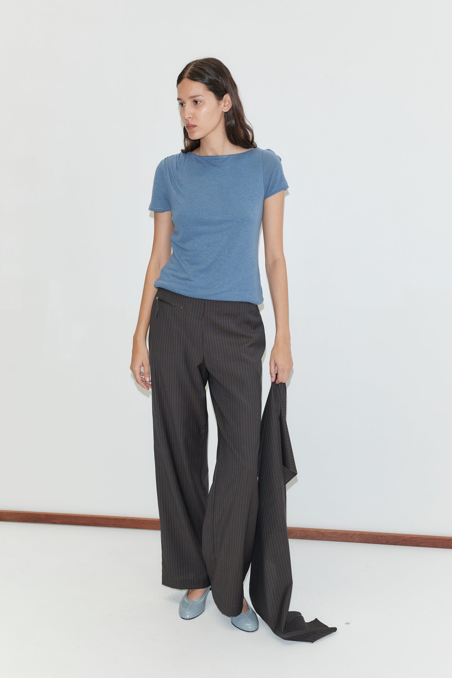A woman stands against a white wall, wearing a blue t-shirt, Pure Wool Zip Pant - Charcoal Stripe, light blue shoes, and holding matching pinstriped fabric in her left hand.