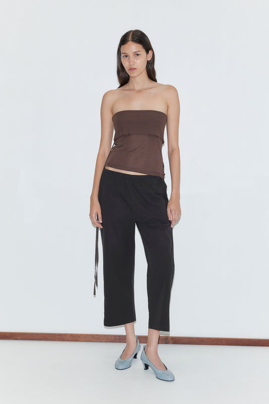 A woman with long dark hair stands against a white background, wearing a brown strapless top, Capri Pant - Black by Deiji Studios, and light blue heels. She looks straight ahead, expression neutral.
