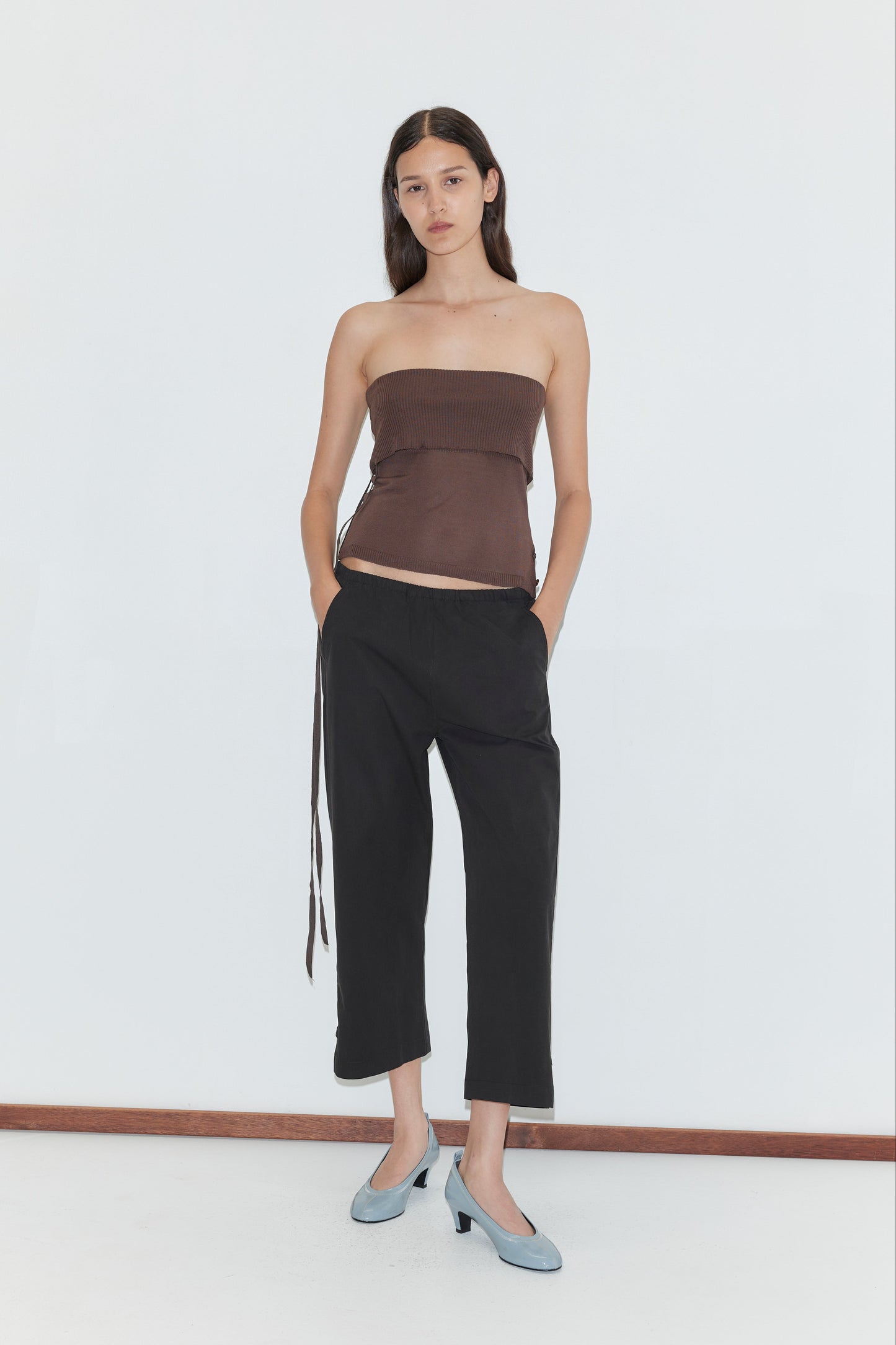 A woman stands against a plain white background wearing a brown strapless top, the Capri Pant in Black from Deiji Studios, and light blue low-heeled shoes. Her hands are in her pockets and her expression is neutral.