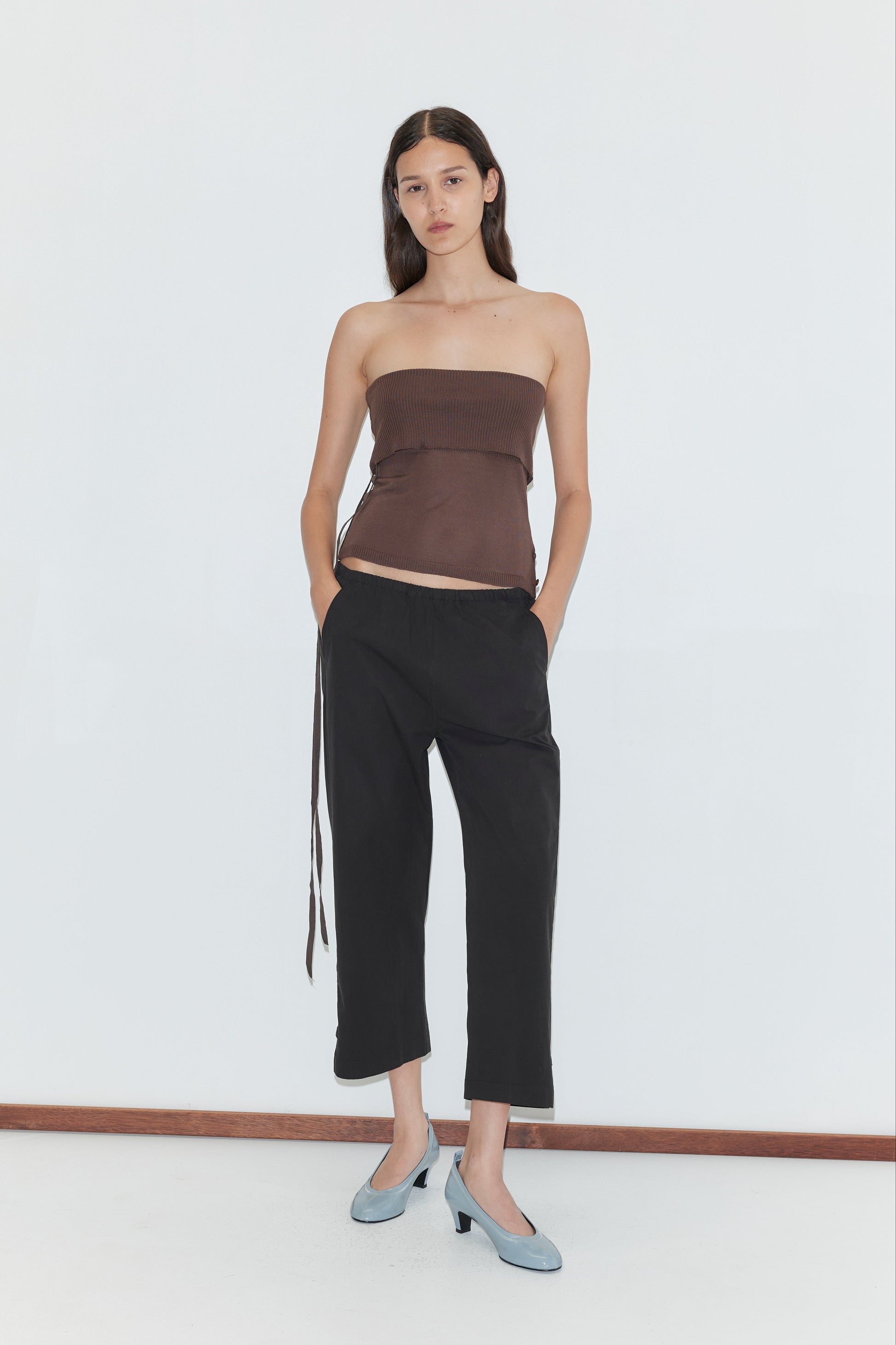 A woman stands against a plain white background wearing a brown strapless top, the Capri Pant in Black from Deiji Studios, and light blue low-heeled shoes. Her hands are in her pockets and her expression is neutral.
