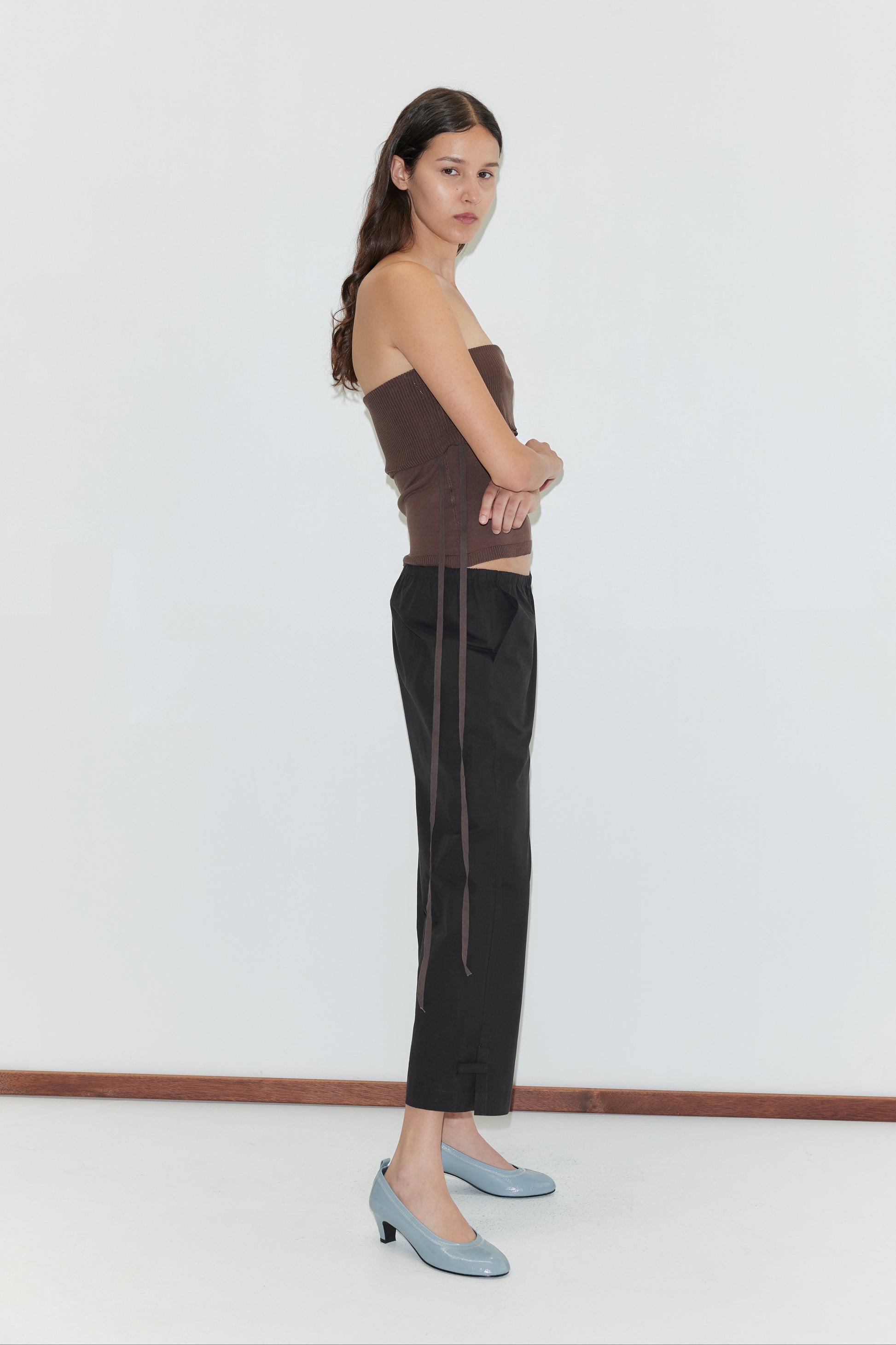 A woman with long dark hair stands sideways against a white wall, arms crossed, wearing a brown strapless top, Capri Pant - Black from Deiji Studios, and light blue heeled shoes.