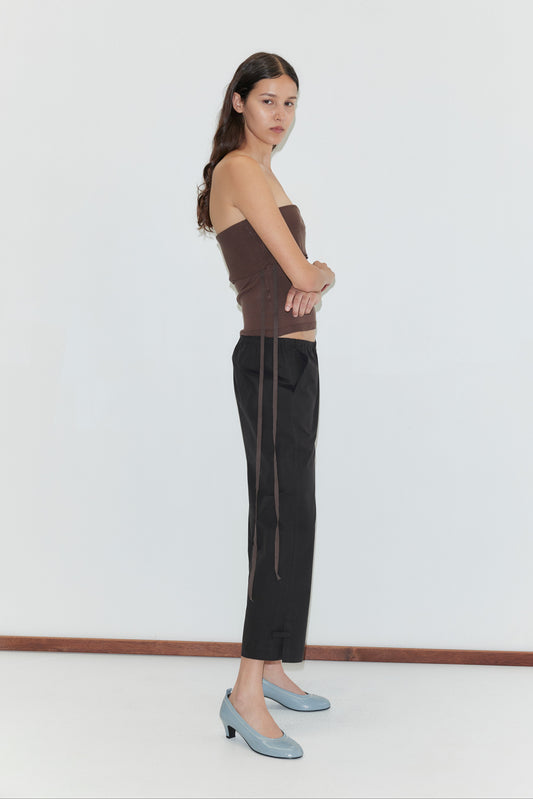 A woman with long dark hair stands sideways against a white wall, arms crossed, wearing a brown strapless top, Capri Pant - Black from Deiji Studios, and light blue heeled shoes.