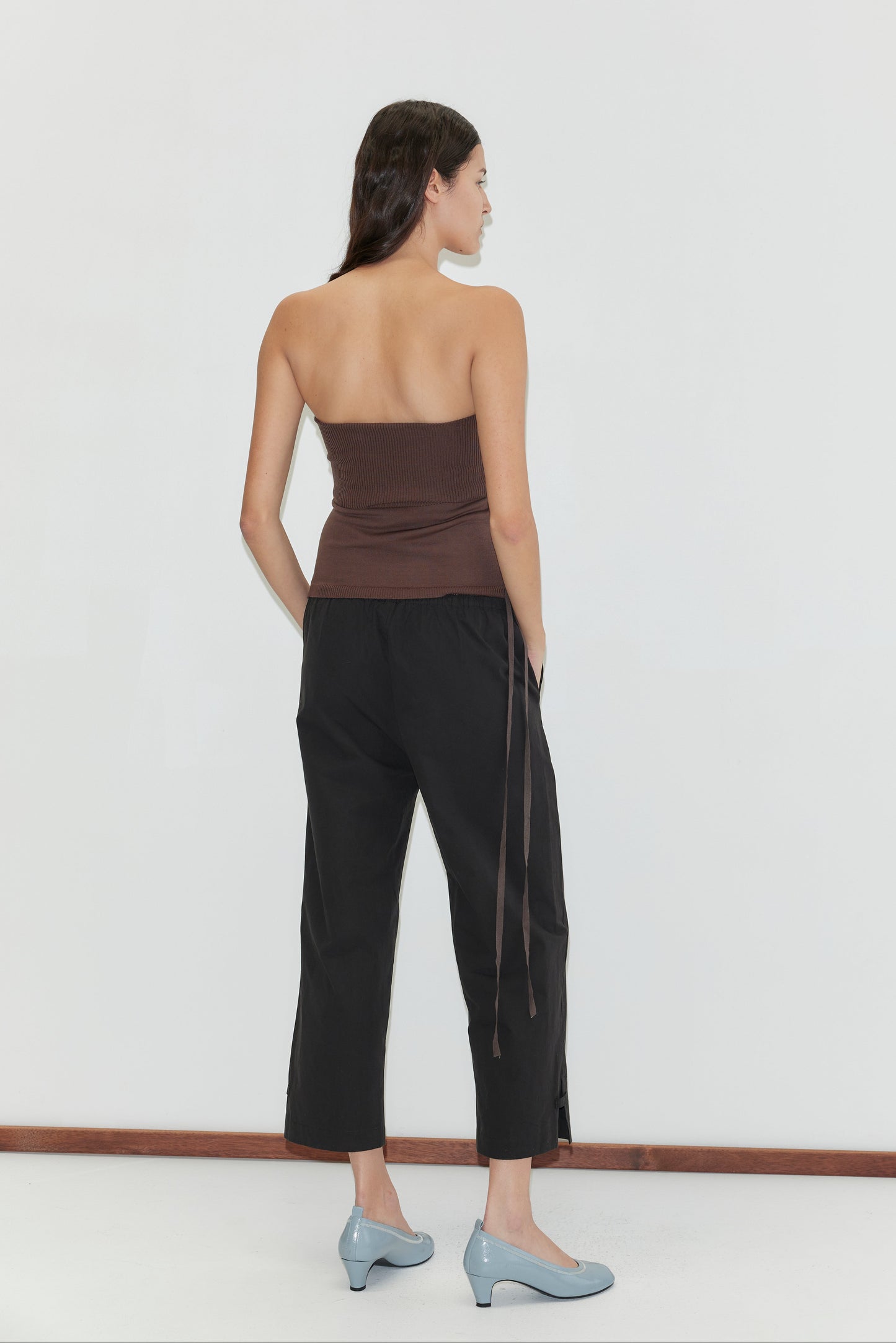 A woman stands with her back to the camera against a white background, wearing a brown strapless top, Capri Pant in black, and light blue low-heeled shoes.