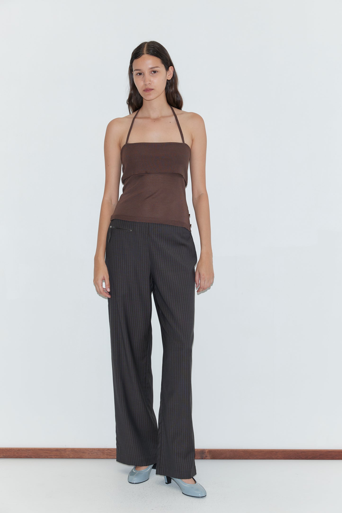 A woman with long dark hair and a neutral expression wears the Pure Wool Zip Pant - Charcoal Stripe, a brown halter top, and light blue heels against a plain white background.