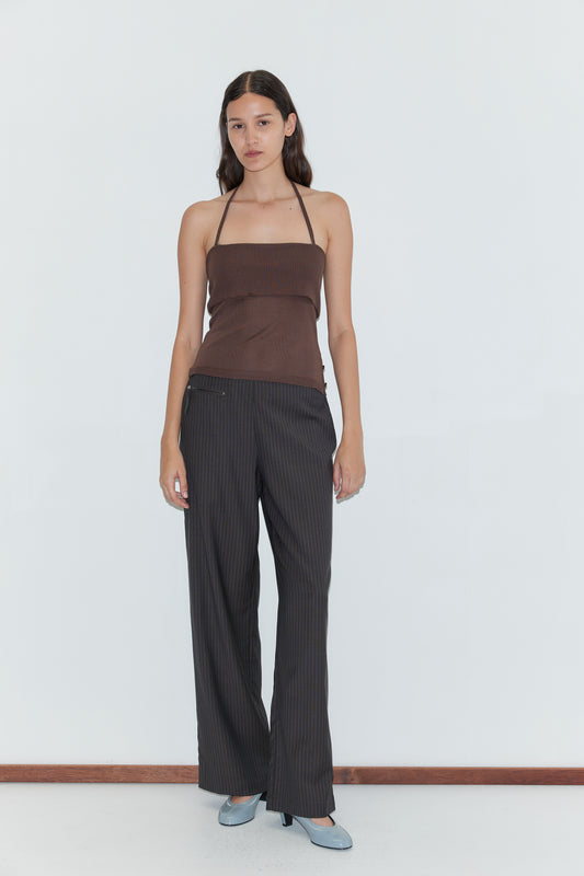 A woman with long dark hair and a neutral expression wears the Pure Wool Zip Pant - Charcoal Stripe, a brown halter top, and light blue heels against a plain white background.