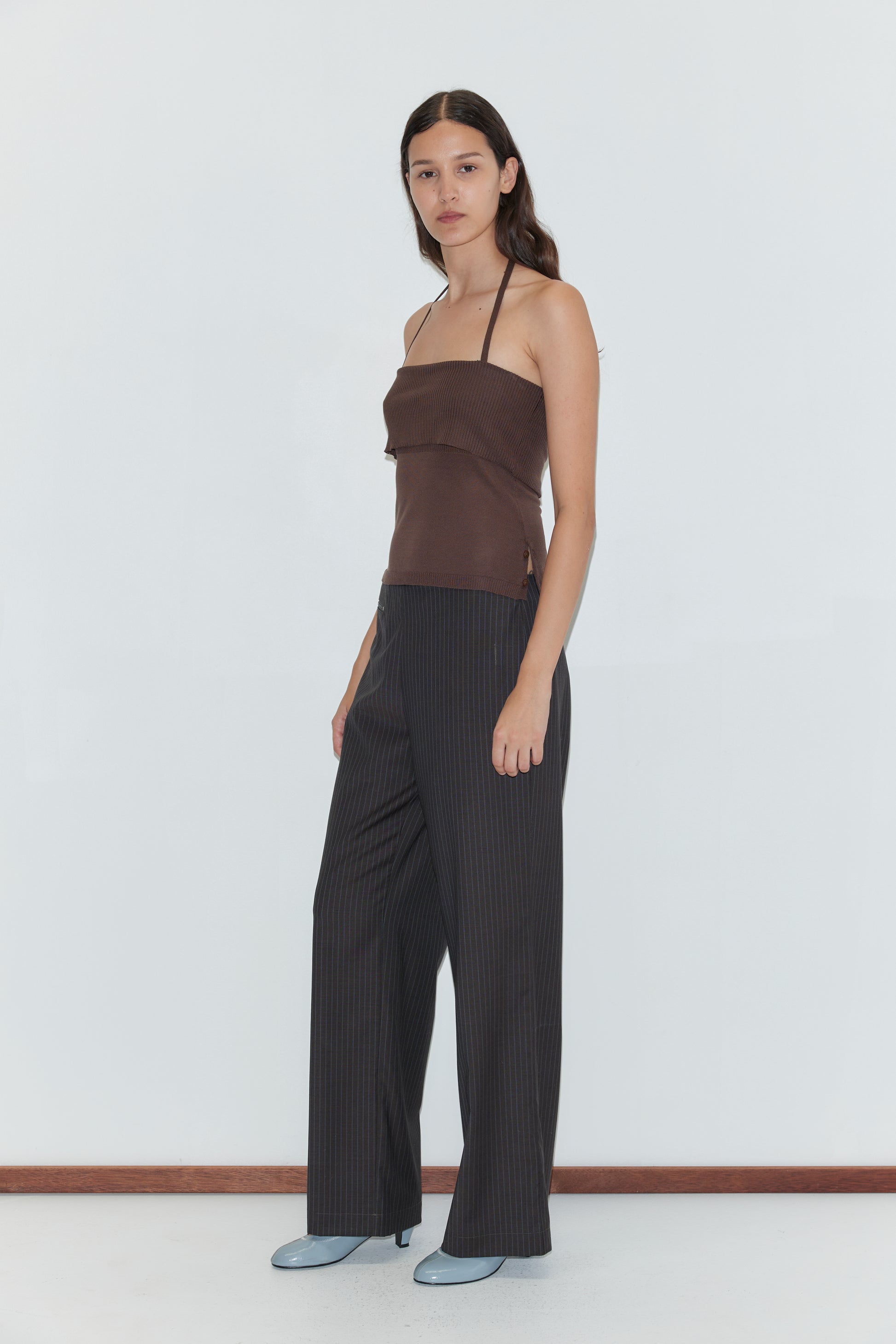 A woman with long dark hair stands in a brown halter top and the Pure Wool Zip Pant - Charcoal Stripe, paired with light blue shoes, against a plain white background. She faces slightly to the side with a neutral expression.