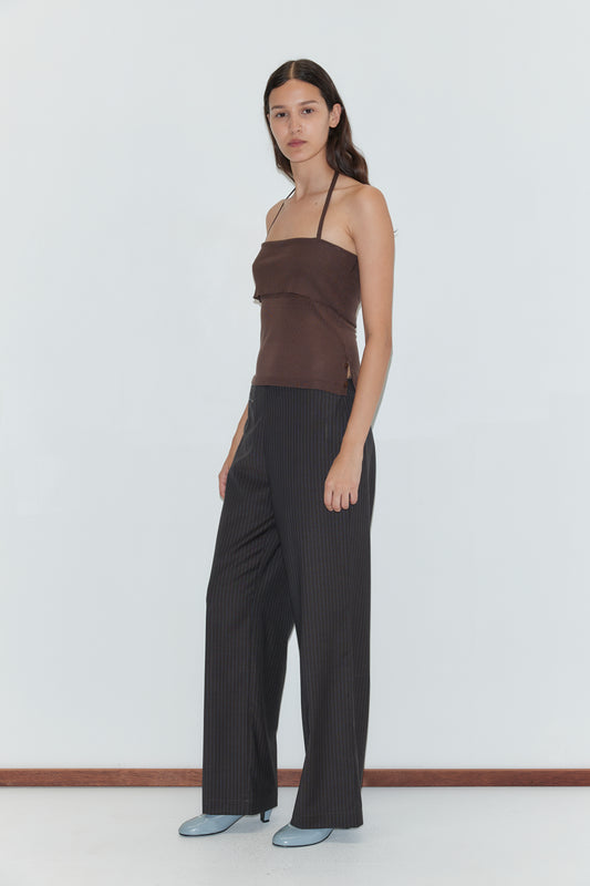 A woman with long dark hair stands in a brown halter top and the Pure Wool Zip Pant - Charcoal Stripe, paired with light blue shoes, against a plain white background. She faces slightly to the side with a neutral expression.