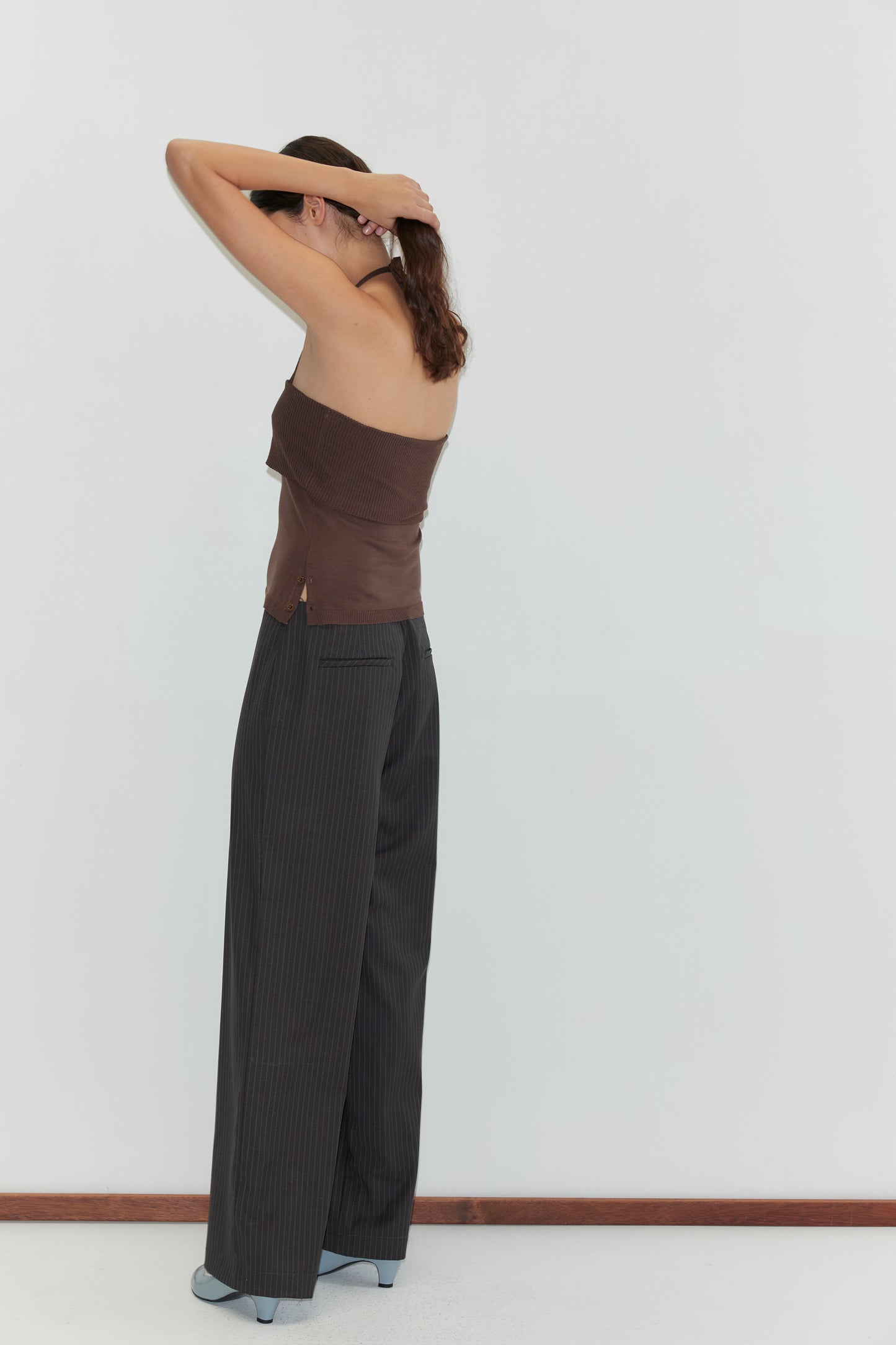 A woman stands against a plain white wall, facing away with raised arms as she adjusts her hair. She wears a brown strapless top, Pure Wool Zip Pant - Charcoal Stripe with relaxed legs, and gray shoes.
