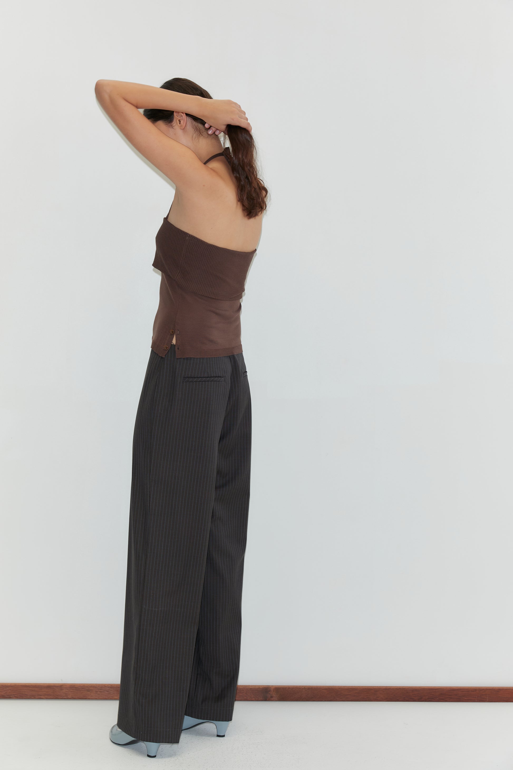 A woman stands against a plain white wall, facing away with raised arms as she adjusts her hair. She wears a brown strapless top, Pure Wool Zip Pant - Charcoal Stripe with relaxed legs, and gray shoes.