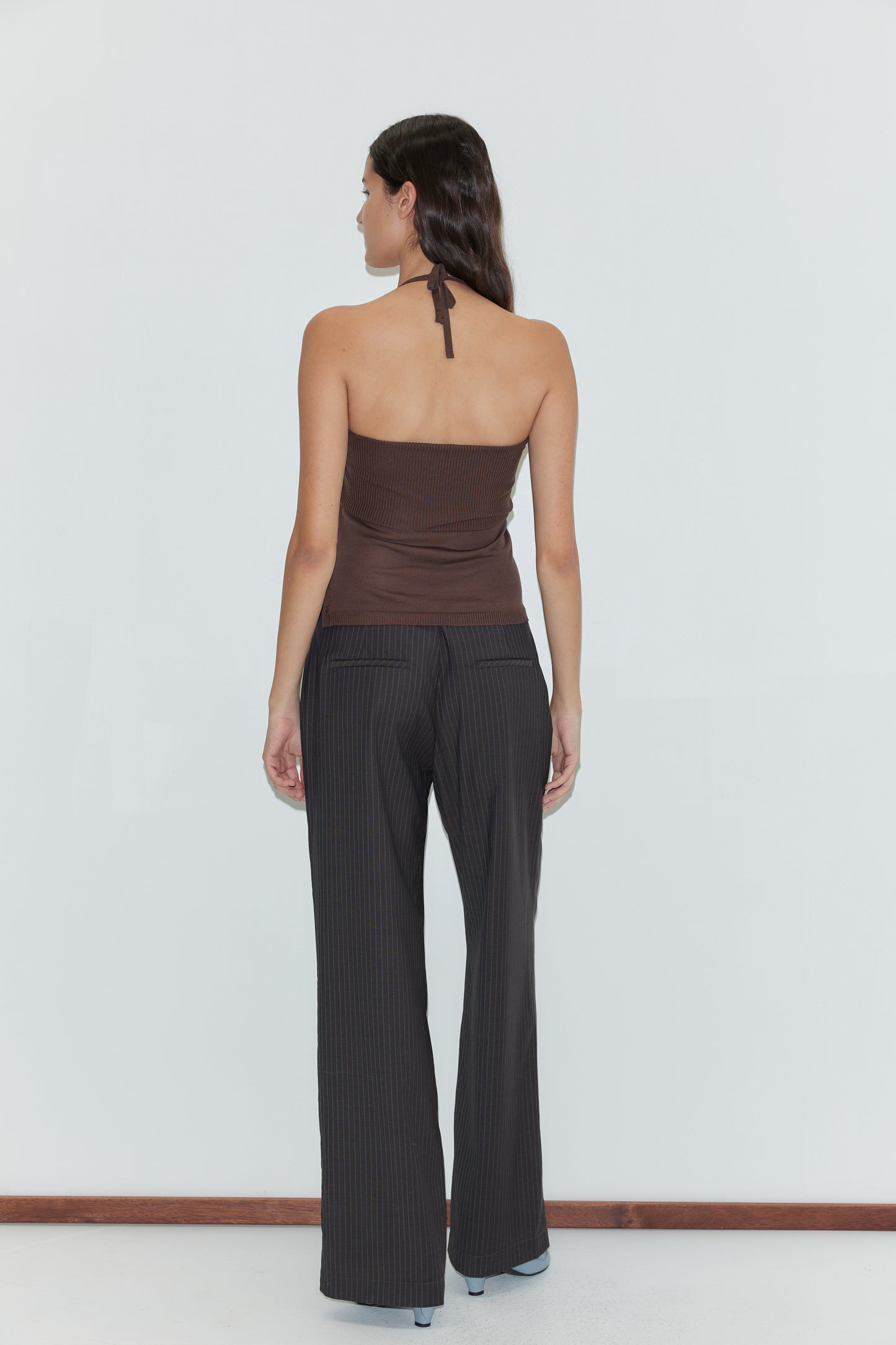 A woman with long dark hair stands facing away from the camera in front of a plain light background, wearing a brown halter top and the Pure Wool Zip Pant - Charcoal Stripe.