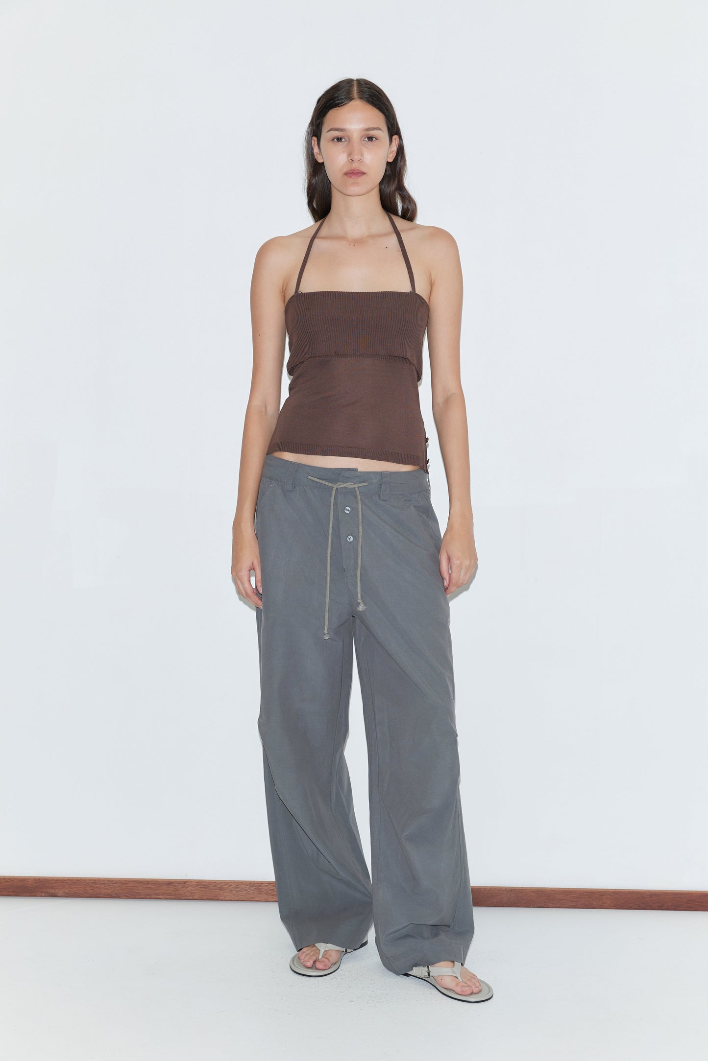 A person stands against a white background wearing a brown strapless top, flip-flops, and the Deiji Studios Panel Leg Pant in Charcoal, which features a relaxed fit and drawstring waist. They have long, straight hair and a neutral expression.