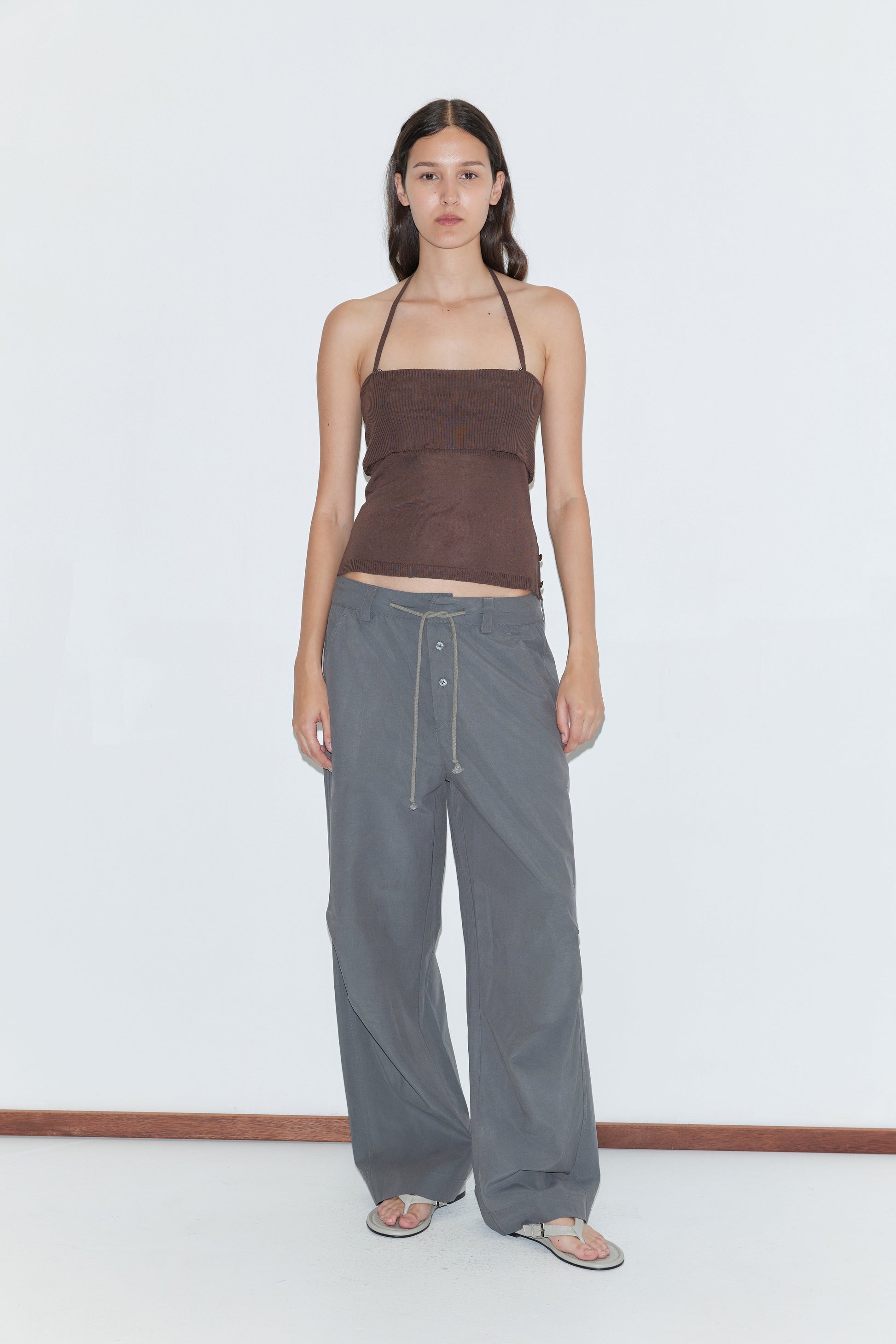 A person stands against a white background wearing a brown strapless top, flip-flops, and the Deiji Studios Panel Leg Pant in Charcoal, which features a relaxed fit and drawstring waist. They have long, straight hair and a neutral expression.