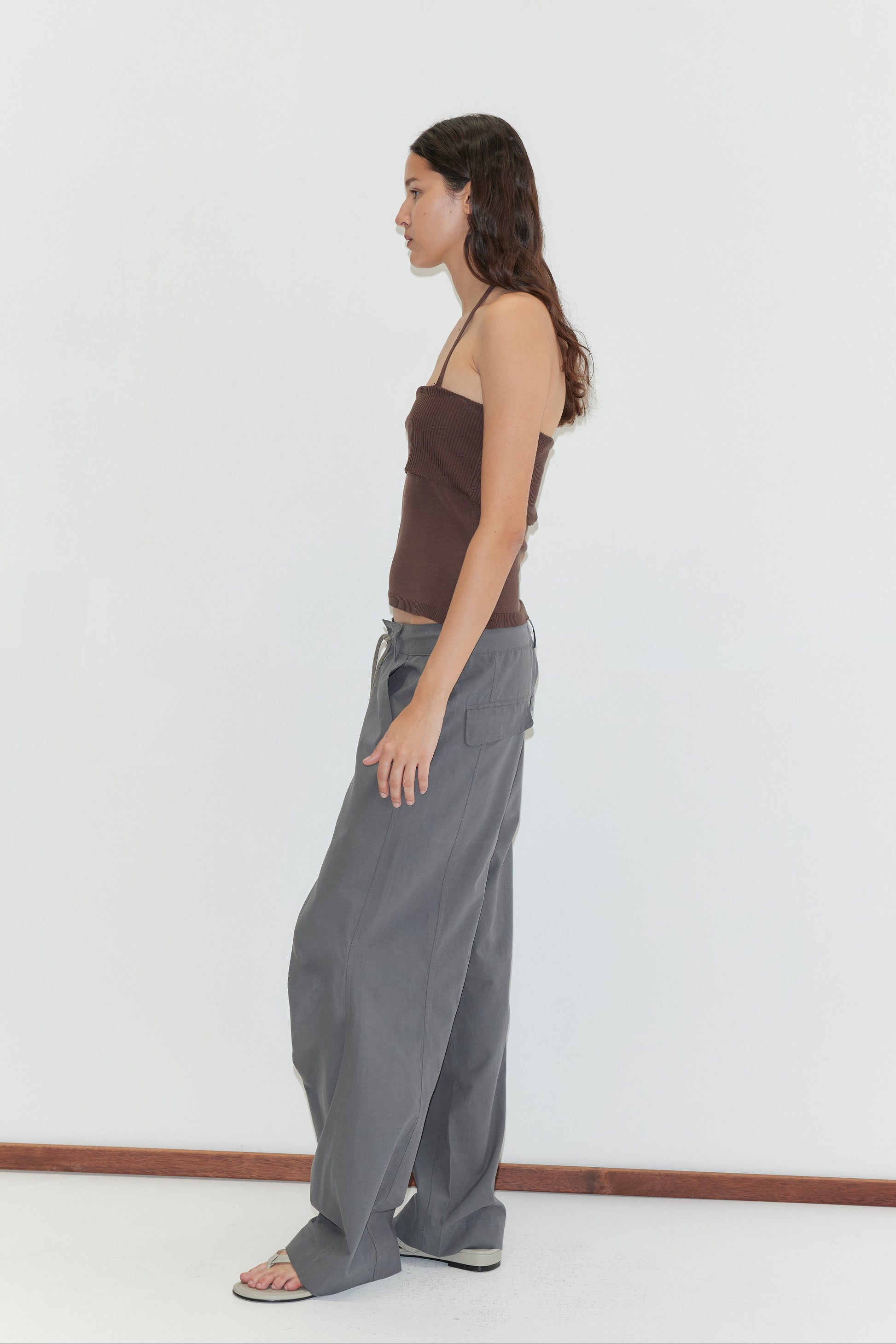 A woman with long brown hair wears a brown strapless top, the Panel Leg Pant in Charcoal from Deiji Studios, and sandals, standing sideways against a white background.