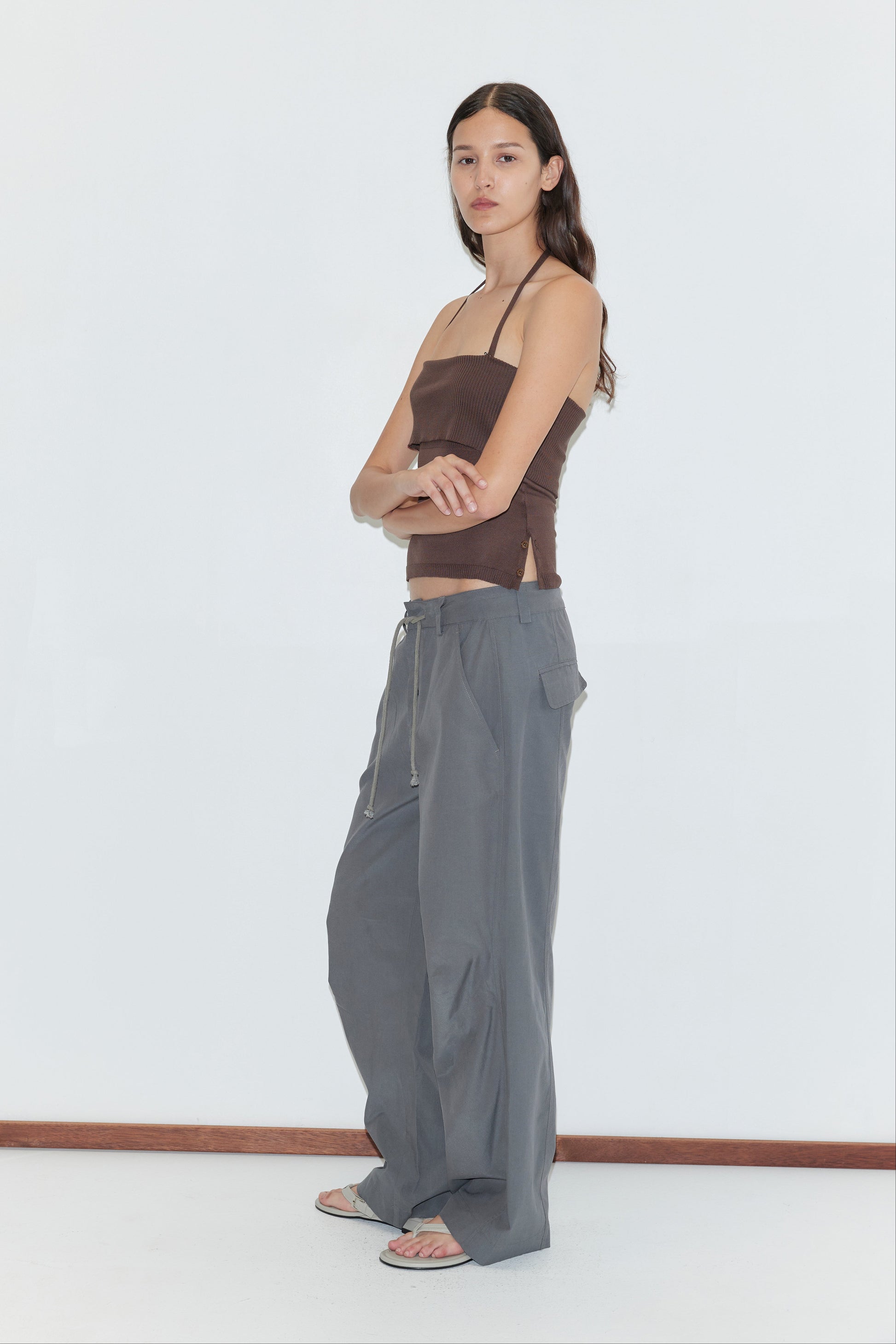 A woman stands sideways wearing a brown strapless top, the Panel Leg Pant in Charcoal by Deiji Studios, and sandals against a plain white background.