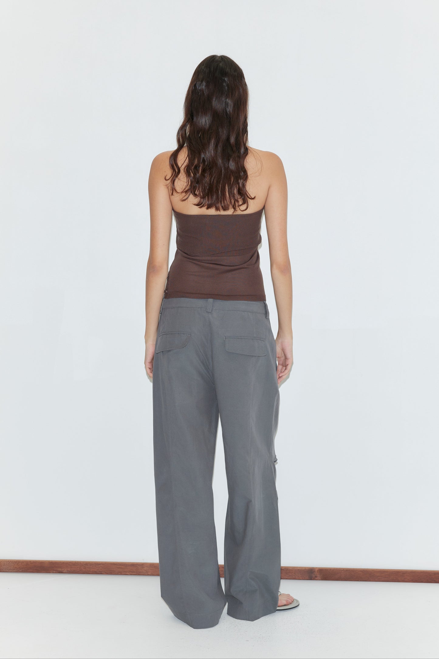 A person with long brown hair stands facing away, wearing a brown strapless top and Panel Leg Pant - Charcoal from Deiji Studios, loose-fitting wide-leg pants, against a plain white background.