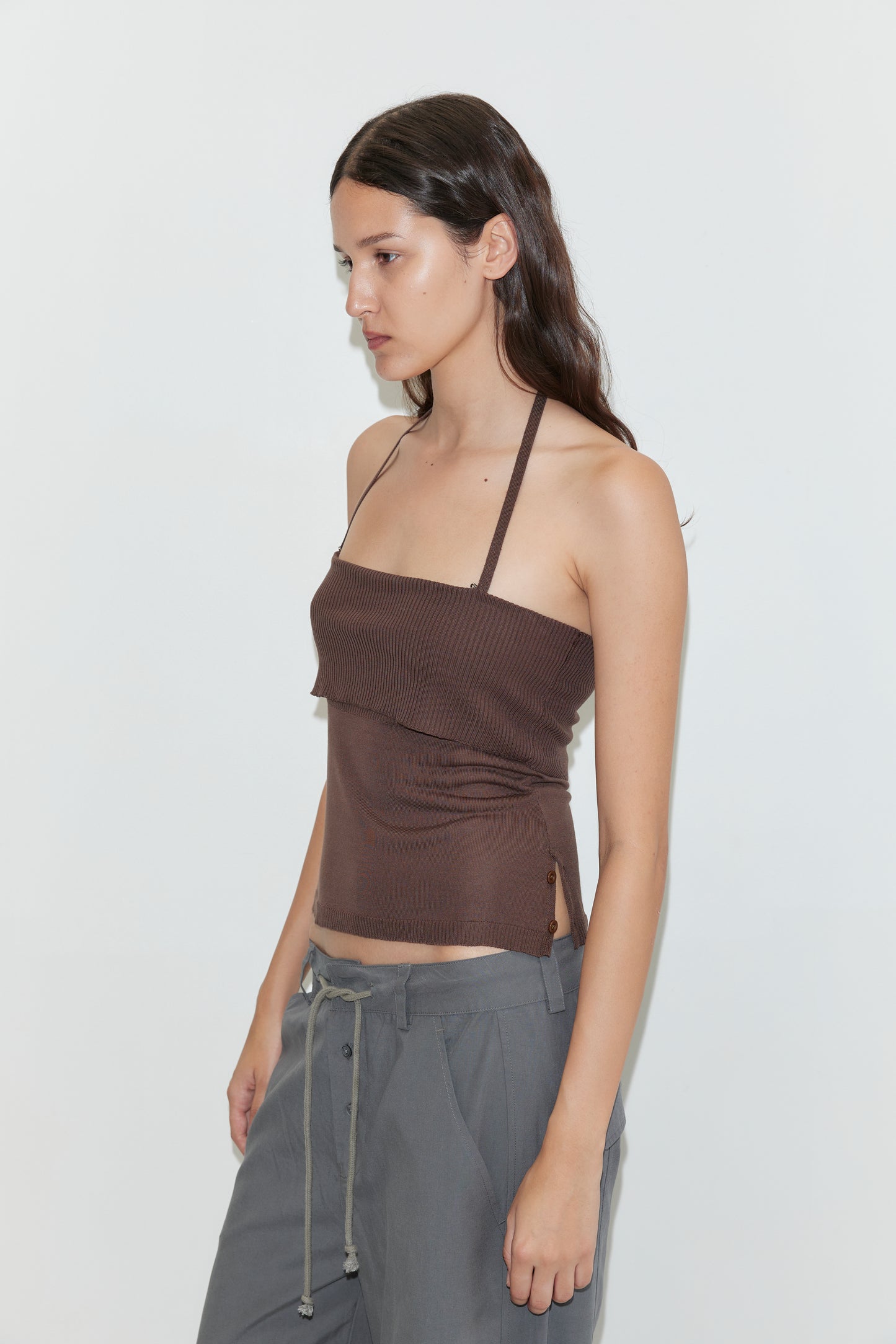 A woman with long brown hair, wearing a brown halter top and Deiji Studios Panel Leg Pant in Charcoal, stands against a plain white background facing left in a relaxed pose.